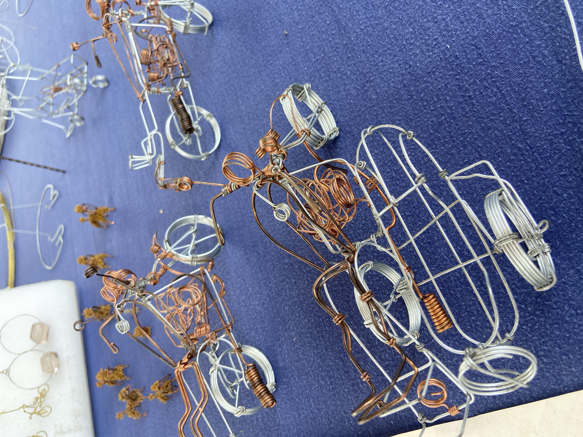 The image shows three wire-frame vehicles on a blue fabric surface. The vehicles are primarily constructed of copper and silver-colored wire. Two of the vehicles appear to be motorcycles, while the third may be a car or another type of motorcycle. The vehicles have wire wheels, and the motorcycle models have coiled-wire components resembling shocks or springs. Some scattered small, brown plant matter or detritus is visible near one of the motorcycles. A white object is partially visible at the lower left of the image, near the same motorcycle. The location is Tinaquillo, Venezuela. There are no people, subjects, or interactions visible.
