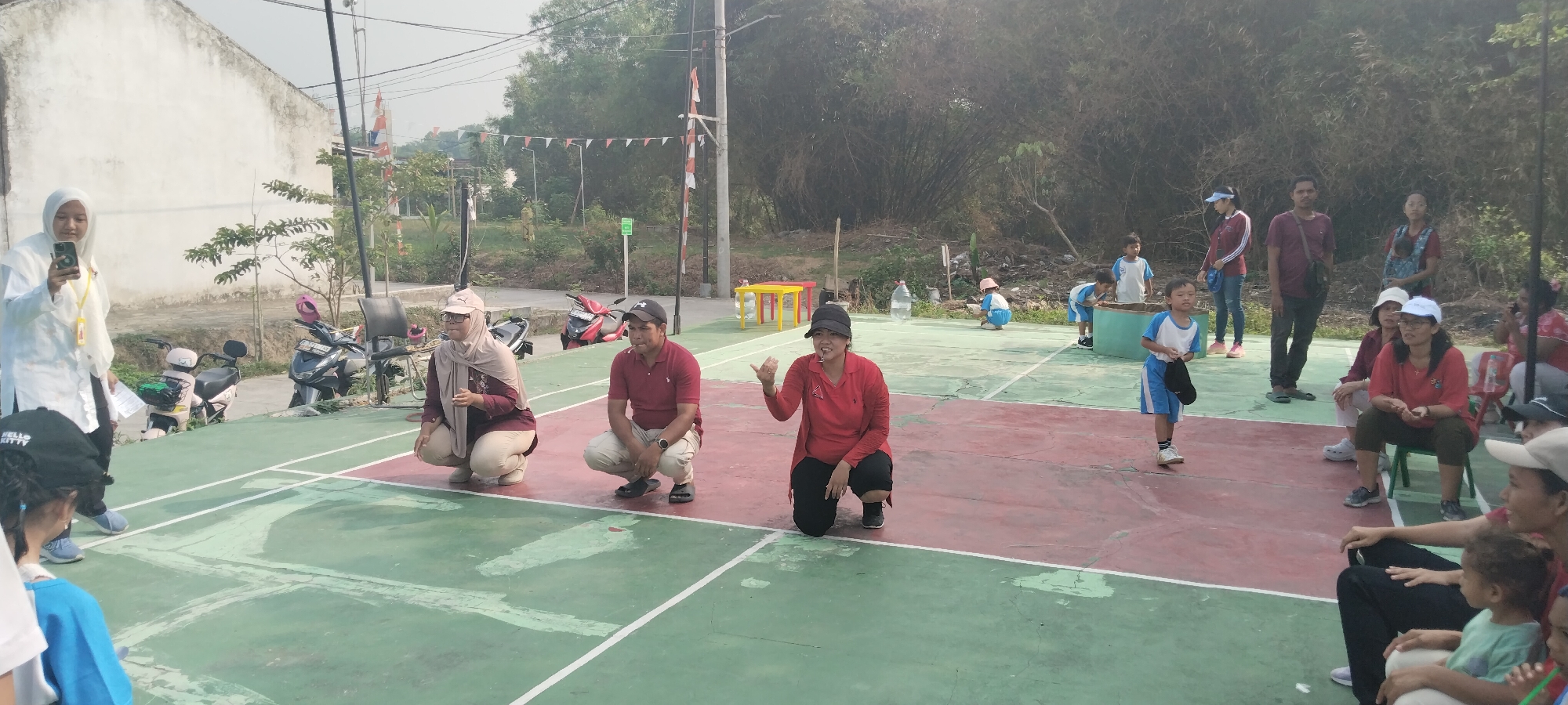 The image depicts an outdoor sports court, likely a badminton court, with visible boundary lines. In the foreground, a woman in a white abaya holds a smartphone and appears to be recording or giving instructions. Several individuals, including adults and children, are present on and around the court. Some adults are crouched down, while others stand or are seated on chairs. Children in athletic wear are visible, some participating in activities. In the background, there are trees, a wall, motorcycles, and other elements suggesting an urban or semi-urban environment. The lighting suggests it is daytime. The presence of Indonesian flags and banners indicates a local event or celebration. The overall scene appears to be a community gathering or sporting event.