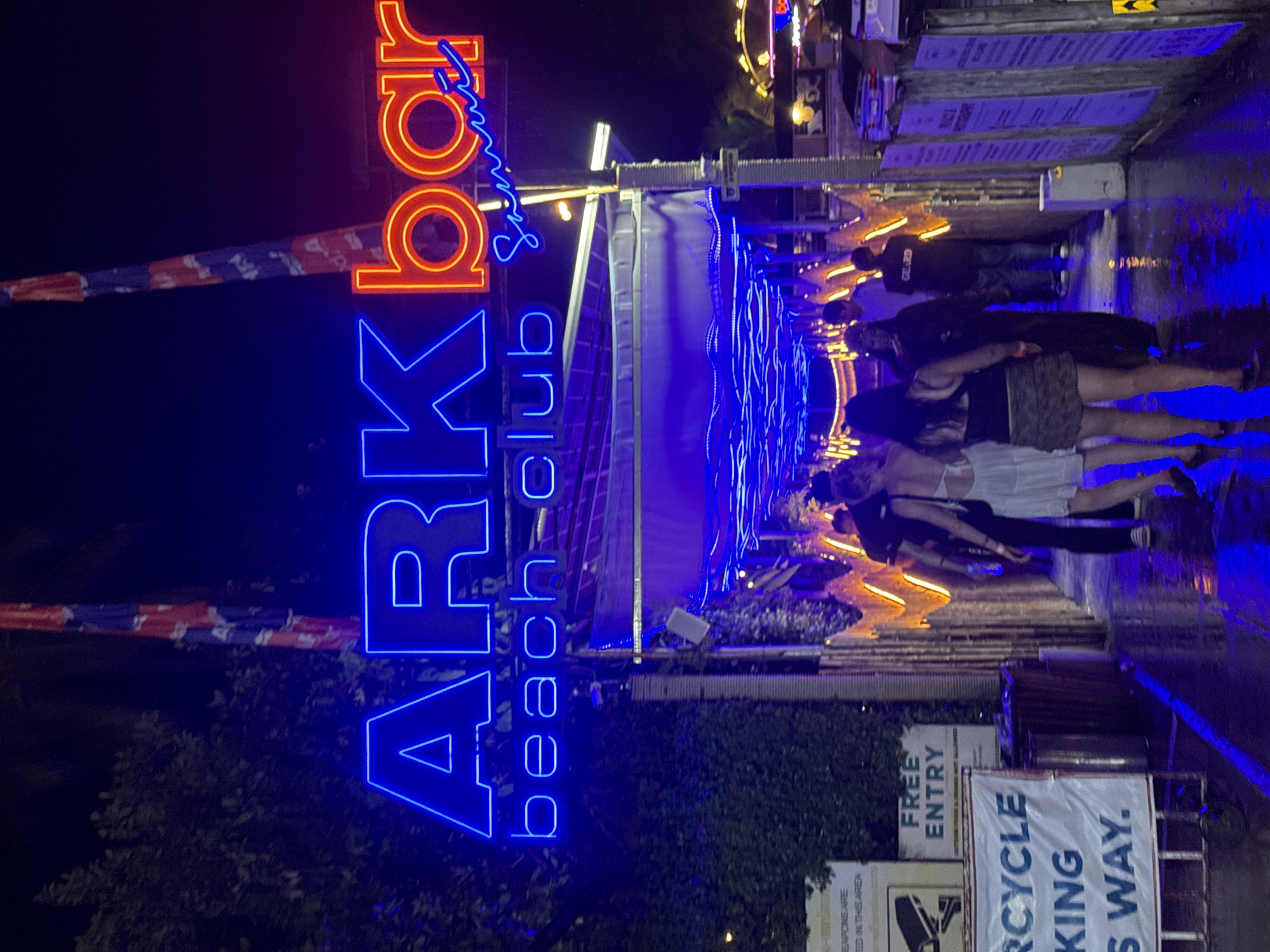 The image shows the entrance to a neon-lit beach club at night, likely in Baan Chaweng, Thailand. A large, brightly lit sign reads "ARK bar beach club Samui" in blue and red neon letters. The entrance walkway is illuminated with blue string lights, and several people are walking through it. Some are dressed in casual attire, and one individual appears to be a security guard. Banners and signs, including one indicating "FREE ENTRY" and another with a "MOTORCYCLE PARKING" notice, are visible. The ground appears wet, suggesting recent rain. The overall scene depicts a vibrant nighttime entertainment venue.