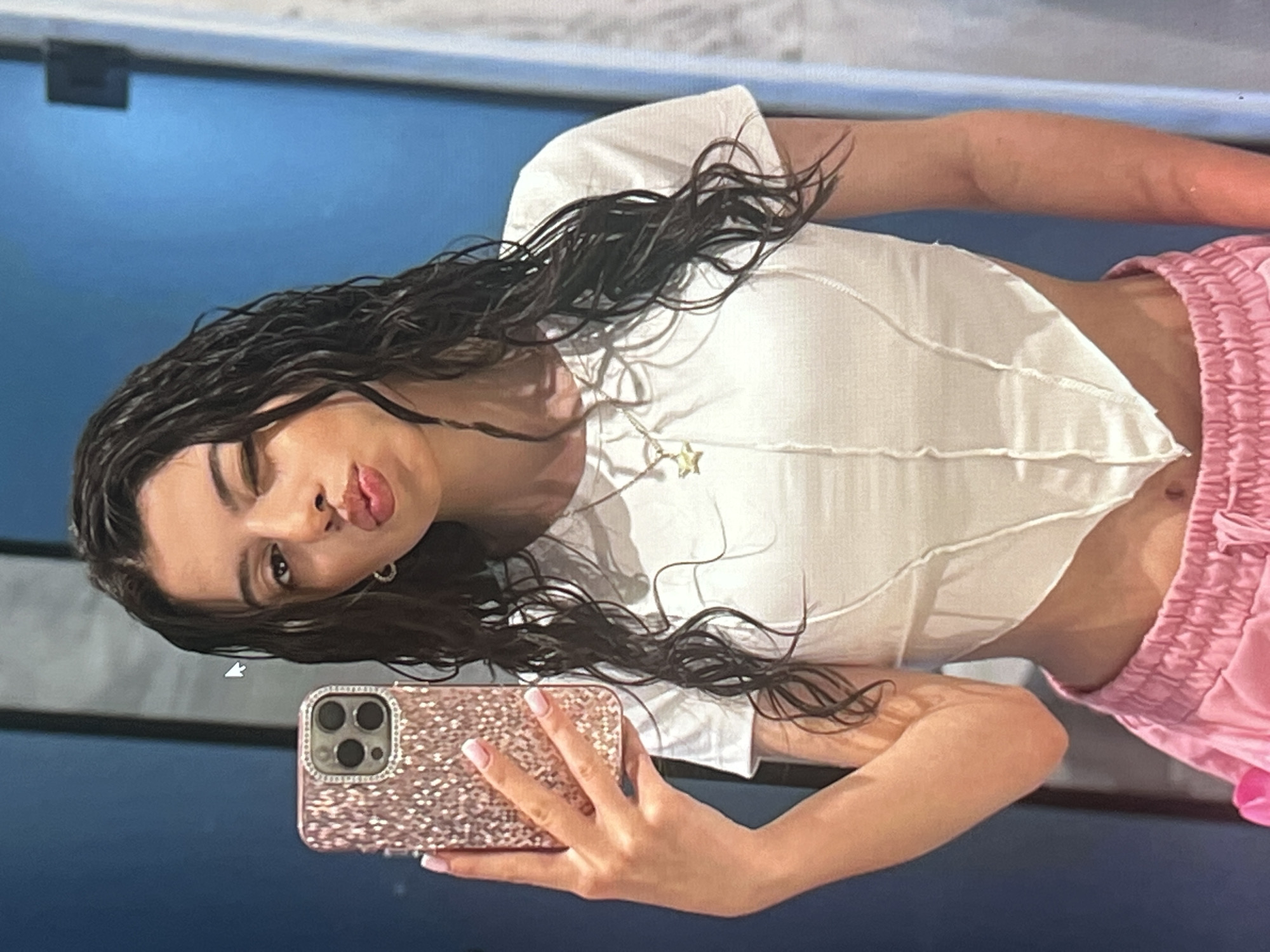 A young woman with long, dark, wavy hair is taking a selfie in a room. She is wearing a white, cropped, ribbed top and pink, ruffled, drawstring shorts. A gold star pendant necklace is visible on her chest. She is winking and puckering her lips, holding a rose gold, glitter-covered phone with a multi-lens camera in her right hand. Her left arm is bent, resting on her hip, and her right arm is extended, holding the phone. The background is out of focus, but appears to be a blue wall. The lighting suggests it is daytime, likely indoors. The overall mood is playful and casual. There is no visible text in the image. The location context is Vanadzor, Armenia.