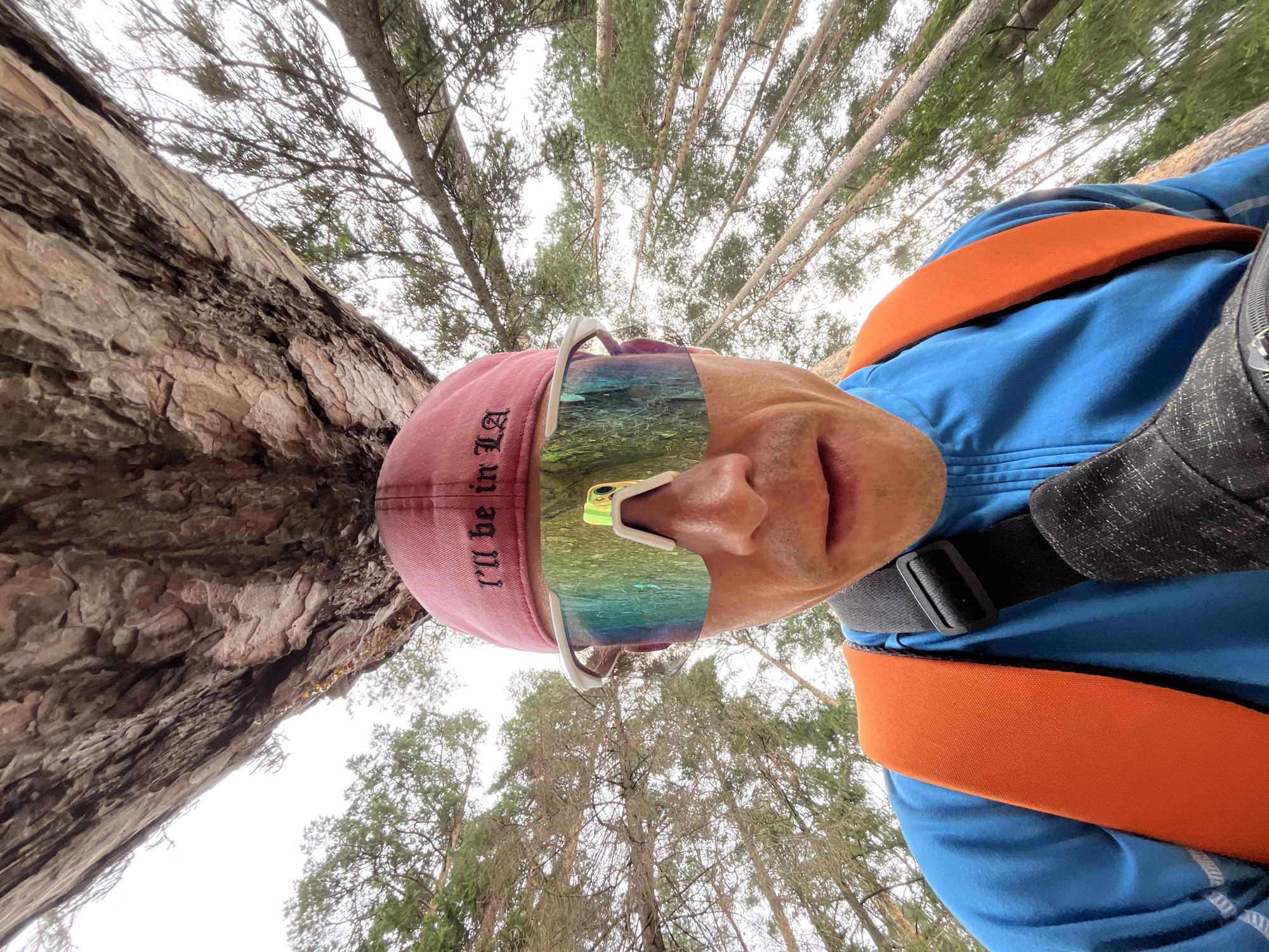 A man wearing a pink baseball cap and mirrored sunglasses is photographed from a low angle looking up at the camera. The cap has text that reads "I'LL BE IN LA". He is wearing a blue jacket and an orange backpack strap is visible across his shoulder. The background consists of tall trees and a cloudy sky. The scene appears to be outdoors, possibly in a forest or park. There is no indication of the city of Chaikovsky, Russia in the image itself.