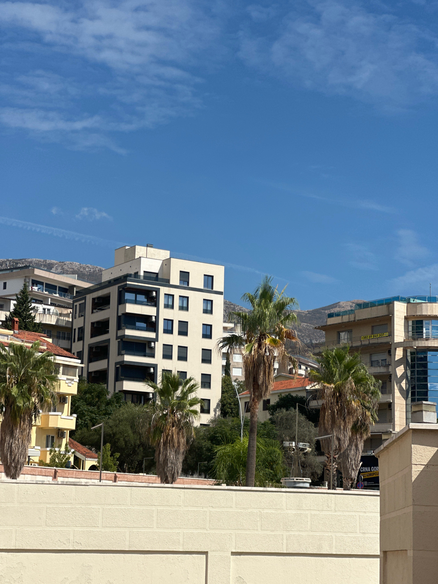 The image depicts an urban scene in Boreti, Montenegro, characterized by a cluster of modern buildings set against a backdrop of hills under a clear blue sky with scattered cirrus clouds. In the foreground, a cream-colored brick wall runs horizontally across the lower portion of the frame. Behind the wall, several palm trees with sparse fronds are visible.
Dominating the mid-ground are several multi-story buildings. One prominent building is a contemporary structure with a light-colored facade, dark window frames, and balconies. To its left, another building features a lighter facade with balconies and is partially obscured by trees. Further to the right, another building has a glass facade and a rooftop terrace. A yellow building with balconies is also present.
In the distance, hills form a natural boundary to the city. The environment suggests a sunny day in a coastal or Mediterranean region, given the presence of palm trees and the architectural styles of the buildings. There are no visible people in the image. The overall impression is one of a developed urban area integrated with natural landscape.
