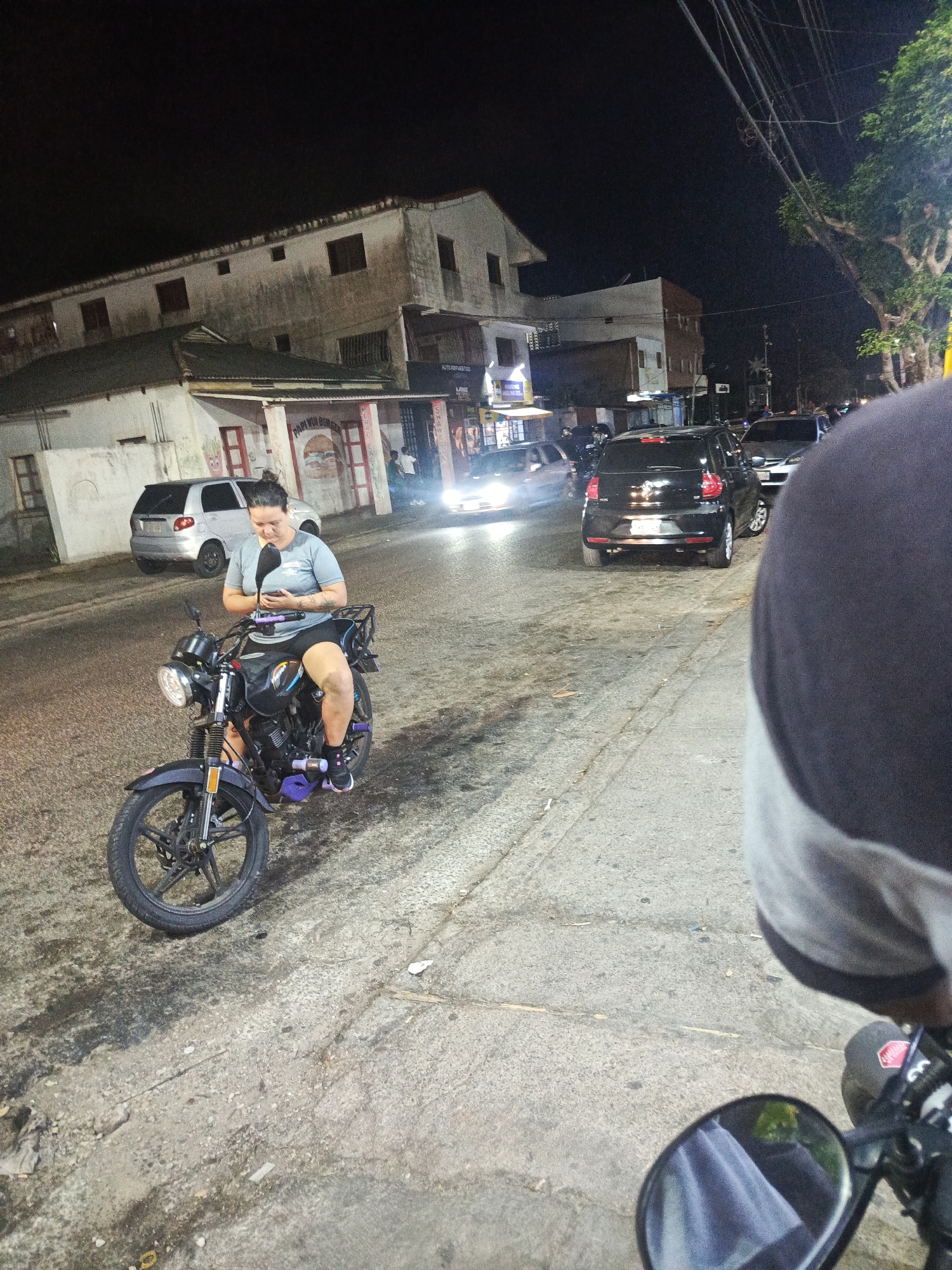 A nighttime street scene in Altagracia, Venezuela, features a woman sitting on a black motorcycle, looking at her phone. She is wearing a gray shirt and black shorts. The motorcycle has purple accents. In the background are parked cars, buildings with lit windows, and streetlights. Part of another person is visible in the foreground to the right. The street is paved and cracked.