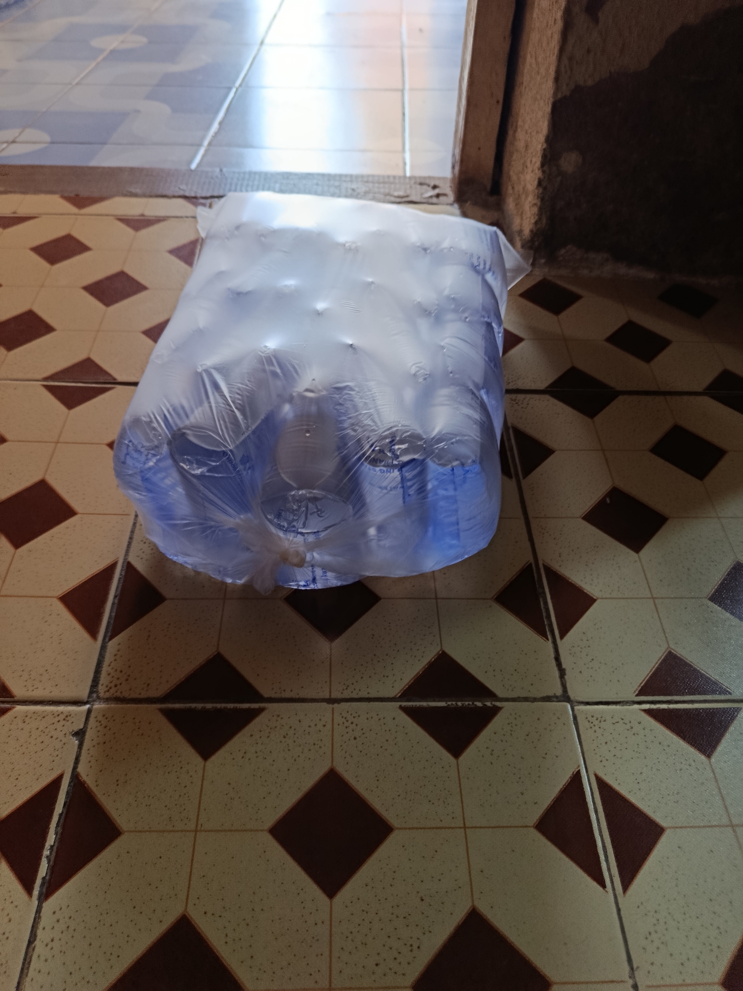 The image shows a plastic-wrapped package of bottled water on a tiled floor. The tiles are a repeating pattern of light beige and dark brown diamonds. The package of water is partially transparent, revealing blue plastic bottles inside. The scene appears to be indoors, possibly a home or a shop, given the presence of tiles and a doorway or opening in the background. The lighting suggests it might be daytime, with light coming from an opening. The overall impression is one of a common household or retail item. There are no people visible in the image. The location of Rafin Par, Nigeria is mentioned in the prompt as context for the scene.