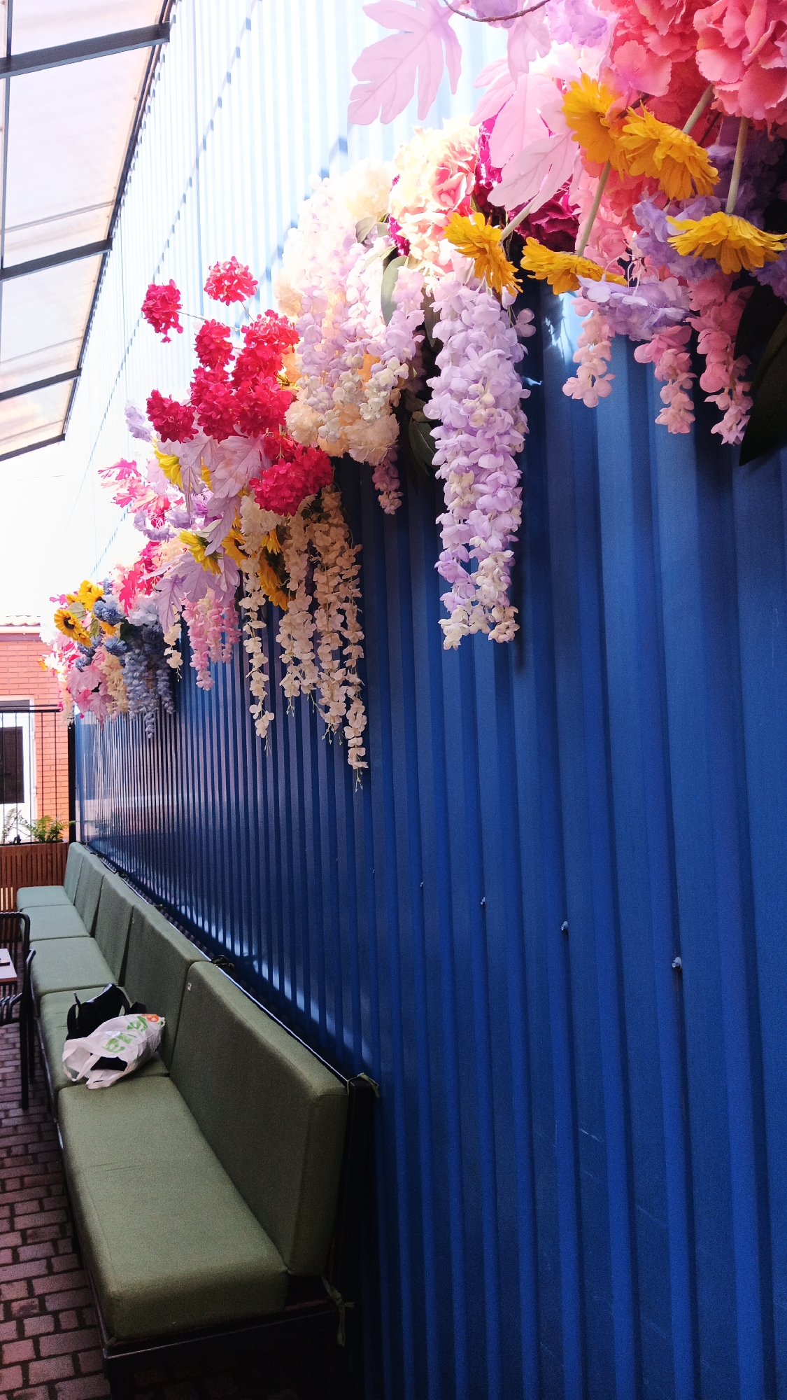 The image depicts an outdoor seating area with a blue corrugated metal wall serving as a backdrop. Along the top of the wall, a colorful display of artificial flowers is arranged, with clusters of pink, yellow, white, and purple blooms cascading downwards. In the foreground, a long, olive-green upholstered bench is positioned along the wall. On the bench, there are two white plastic bags and a black object, possibly a bag or clothing item. The ground beneath the bench is paved with brick. The scene appears to be an exterior of a building, possibly a cafe or restaurant, located in Kryvyi Rih, Ukraine. The lighting suggests it is daytime.