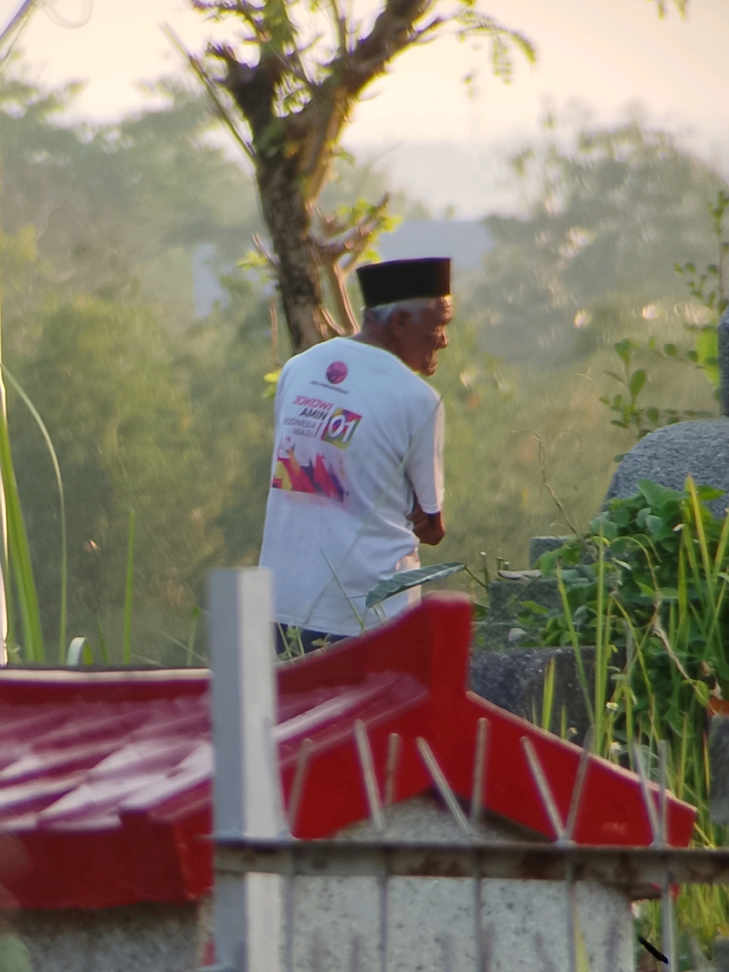 A man wearing a white t-shirt with a black peci cap is standing in an outdoor, slightly out-of-focus environment. The t-shirt has a graphic on the back that includes text and imagery. The man is facing to his right, with his head slightly down. In the foreground, there is a red structure, possibly a roof or enclosure, with vertical poles in front of it. Behind the man, there are trees, greenery, and a stone structure, suggesting a natural or possibly funerary setting. The lighting suggests it is daytime, possibly early morning or late afternoon due to the soft light. There is no indication of the city of Jember, Indonesia, in the image itself.