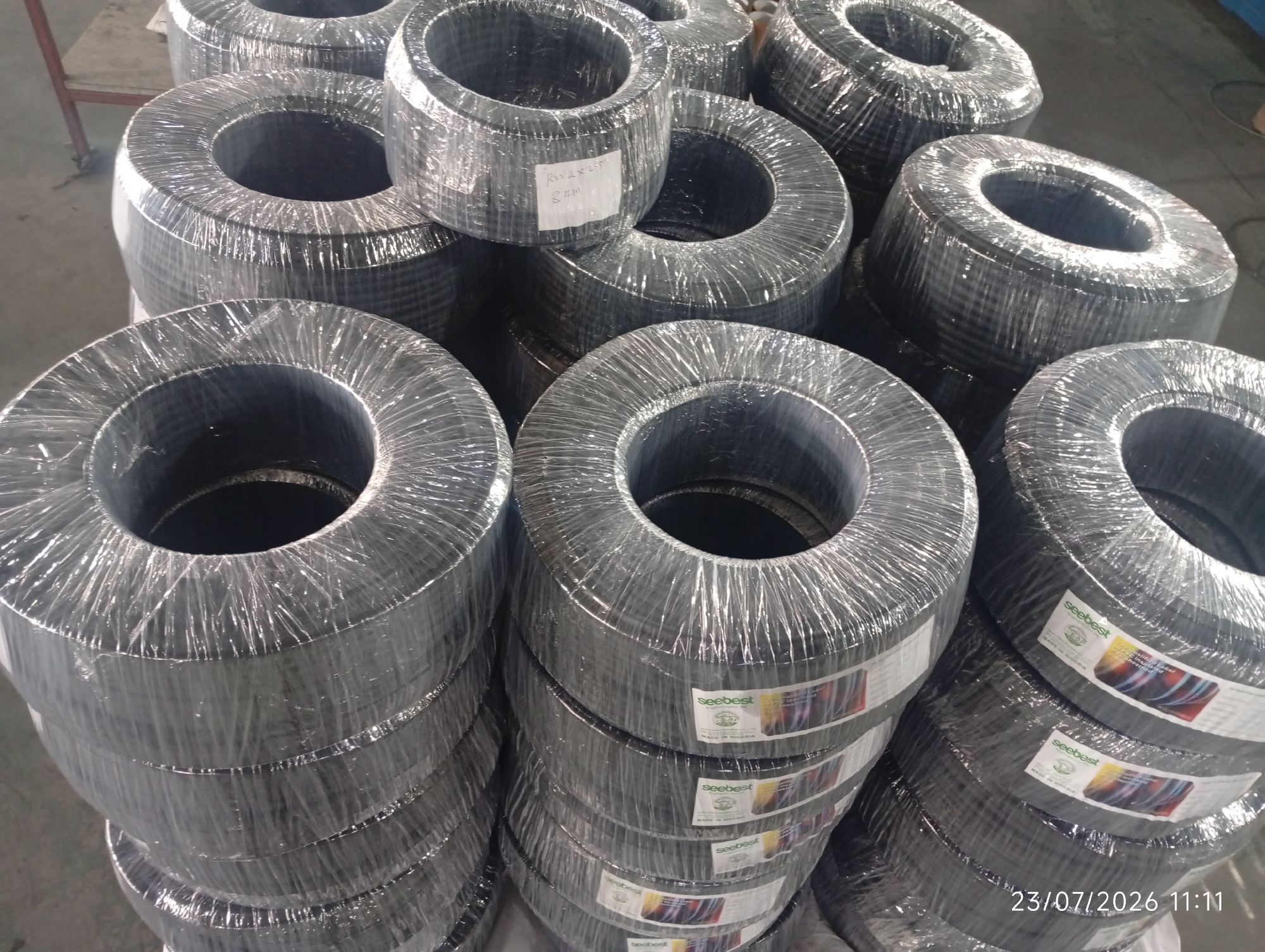 The image shows a warehouse scene in Bana Stream, Nigeria, featuring stacks of coiled electrical cables. The cables are tightly wrapped in clear plastic. Several labels are visible on the plastic wrap, with "SeeBest" being the prominent brand name. One label also indicates "MADE IN NIGERIA". The lighting suggests an indoor setting, possibly a storage facility. The overall impression is one of commercial inventory. A timestamp "23/07/2026 11:11" is overlaid on the bottom right of the image.