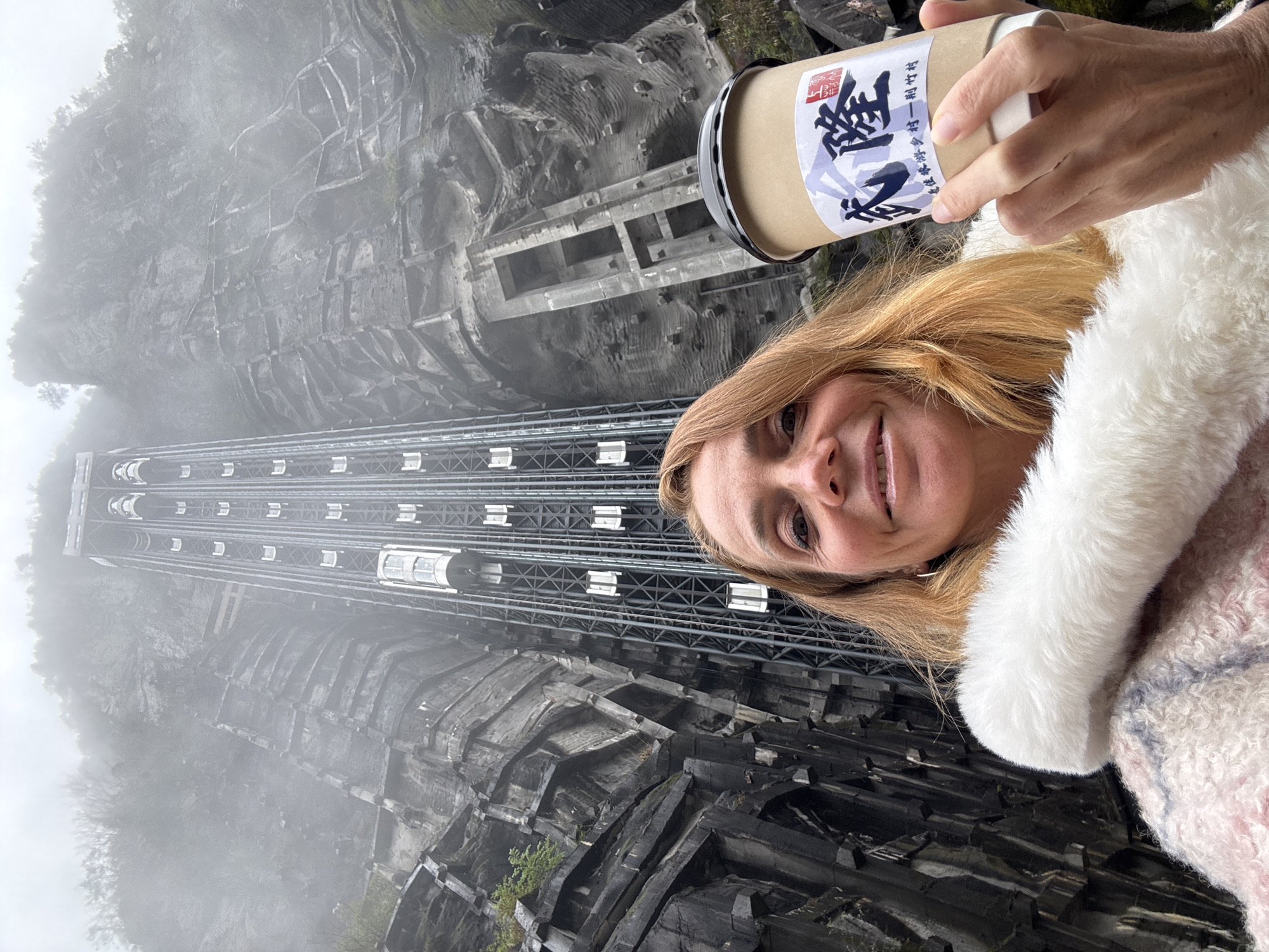 A person, with blonde hair, is holding a beverage container with an Asian language inscription, in front of the Bailong Elevator in Wulong District, China. The elevator is a tall metal structure built into a gray rock face, reaching high into the sky, partially obscured by mist. The person is wearing a furry coat. The elevator is visible in its entirety. The rock face has a textured, layered appearance.