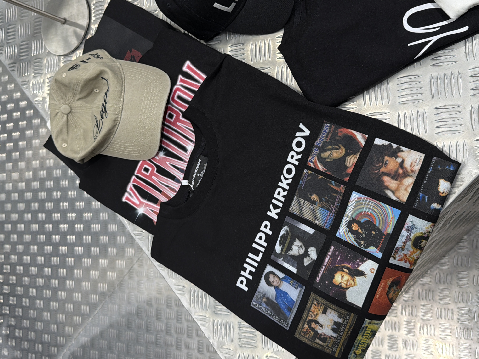The image shows a collection of merchandise, including t-shirts and a baseball cap, laid out on a textured metal surface. The t-shirts are black and feature prominent graphics. One t-shirt displays a large, stylized red and white text that reads "KIRKOROV" and "PHILIPP KIRKOROV". Another t-shirt has a grid of smaller images of the same person, also identified as "PHILIPP KIRKOROV" in white text. Some of these smaller images appear to be album covers or promotional photos, with Cyrillic text overlayed, such as "ФИЛИПП КИРКОРОВ" and song titles. The baseball cap is a light beige color with dark embroidery that includes the word "legend" in a script font and other smaller designs. In the background, a black baseball cap with white lettering is partially visible, along with a glimpse of a metal stand. The overall scene suggests a product display or a close-up view of branded apparel. The metal surface is reflective and appears to be a metallic tread plate, commonly found in industrial or public spaces. The lighting is even, suggesting an indoor setting, likely a retail or exhibition space. The context provided indicates Moscow, Russia, which aligns with the Cyrillic text visible on the merchandise.