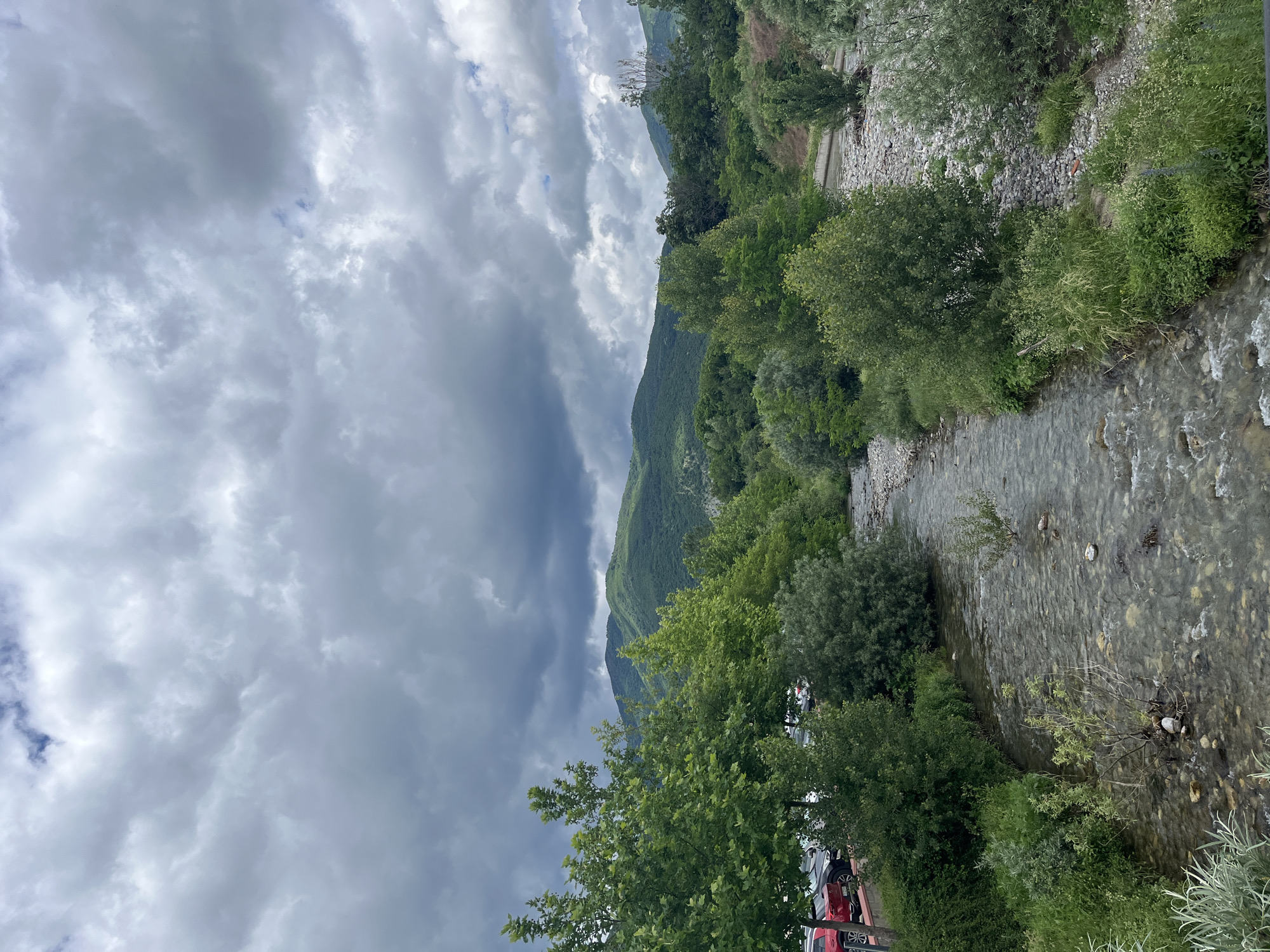 The image depicts a natural landscape near Skobelevo, Bulgaria, under a cloudy sky. A narrow, clear river flows through the foreground, with a rocky bed visible beneath the water's surface. Lush green trees and dense vegetation line the banks of the river, with some trees taller and more prominent than others. In the background, rolling green hills or mountains rise, their peaks partially obscured by the overcast conditions. On the left side of the image, a parking area with several cars is visible, suggesting proximity to a developed area. The overall scene conveys a sense of natural beauty with a touch of human presence.