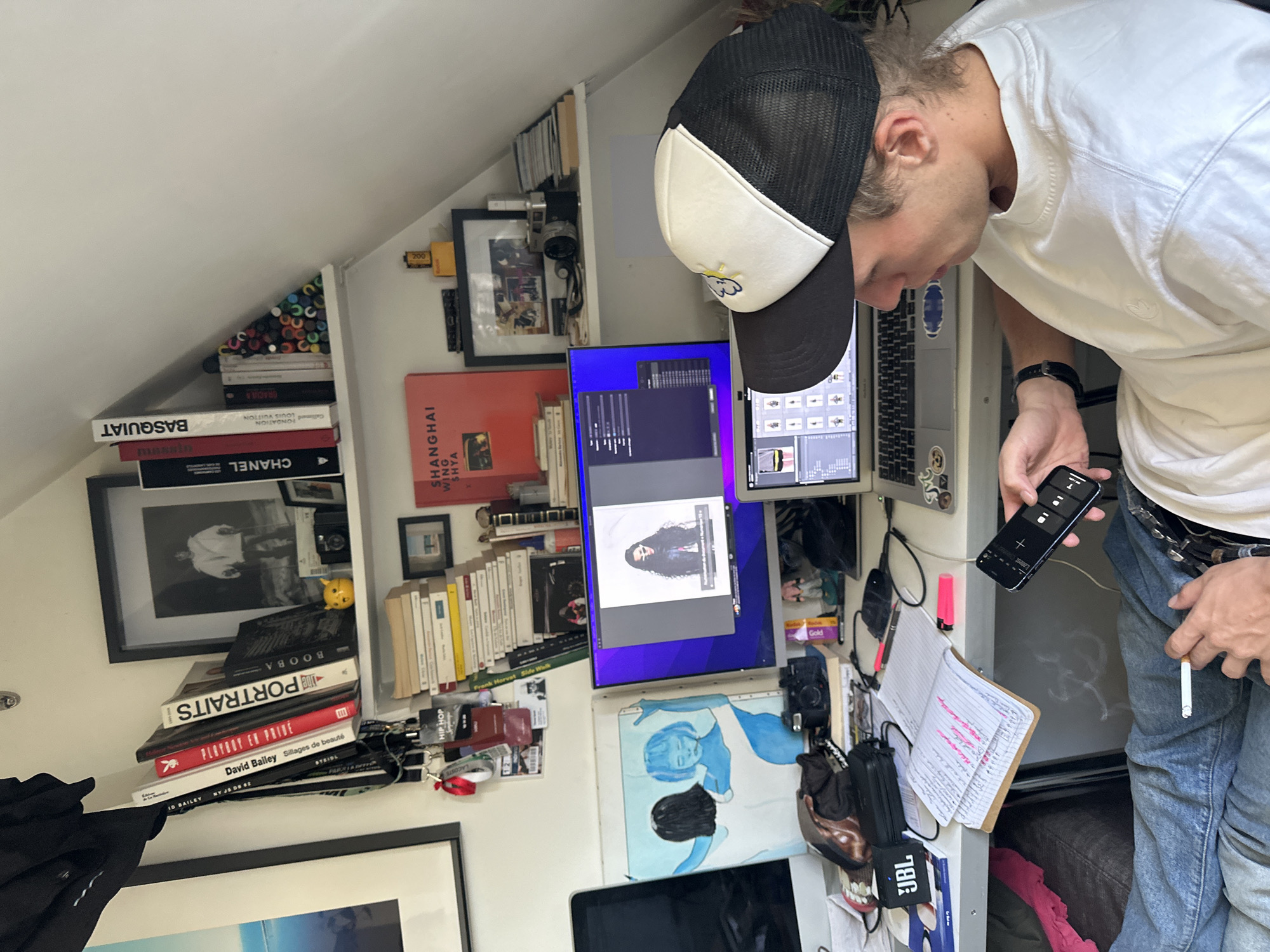 This image depicts a man leaning over a cluttered and personalized workspace, likely a home office in Paris, France. The man, with light brown hair, is wearing a white t-shirt, blue jeans, and a black and white trucker hat. In his left hand, he holds a black smartphone displaying controls, while a lit cigarette is held casually in his right hand.
The workspace is equipped with a dual-monitor setup and a laptop (a MacBook), indicating a focus on digital tasks, possibly photo editing or graphic design, as one vertical monitor displays an image of a woman. The desk and surrounding shelves are densely packed with books, cameras, and various personal items. Visible book titles and subjects suggest interests in art, fashion, and photography, including "BASQUIAT," "CHANEL," "LOUIS VUITTON," "David Bailey," "PORTRAITS," "PLAYBOY EN PRIVÉ," and "SHANGHAI SHYA."
Other notable objects include rolls of Kodak Gold 200 film, a JBL portable speaker, a notebook with handwritten notes and highlights, lanyards, and a small yellow figurine. An abstract artwork in blue and black hangs nearby. The room features a sloped white ceiling, characteristic of an attic or mansard space. The overall impression is that of a creative and busy environment.
Visible text includes: "JBL," "Kodak Gold 200," "BASQUIAT," "CHANEL," "GRACILA," "FONDATION LOUIS VUITTON," "SHANGHAI SHYA," "BOOBA," "PORTRAITS," "PLAYBOY EN PRIVE," "David Bailey," "Sillages de beauté," "NY J5 D8 82," "TEIDL," "MARSIL," "HIP HOP," "LACOSTE," and "Gallimard."
