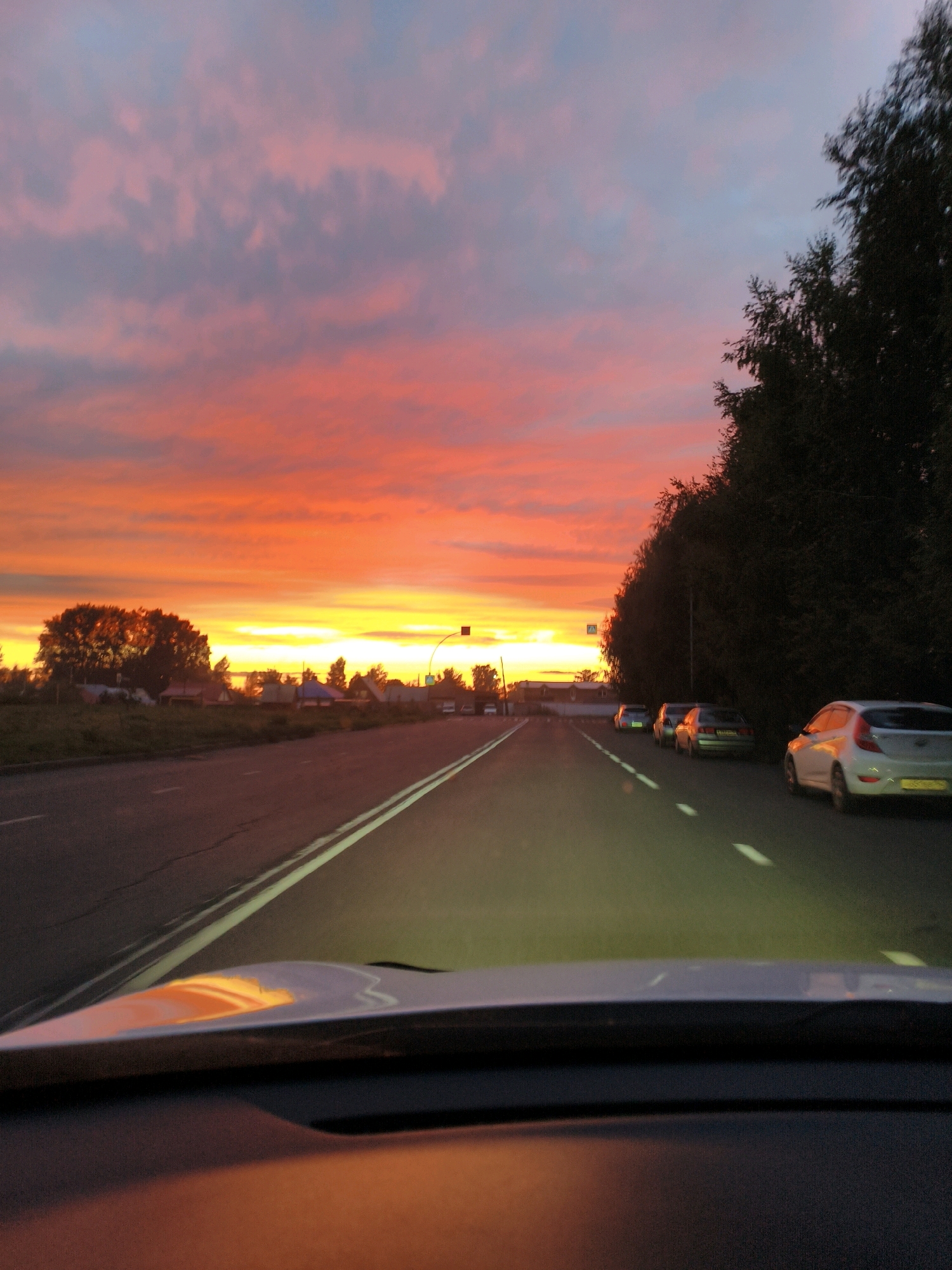 The image captures a sunset scene on a road in the city of Kemerovo, Russia. The sky is ablaze with vibrant hues of orange, pink, and yellow, indicating a dramatic sunset. A road stretches into the distance, with white lane markings visible. Several cars are driving on the road, with one white car in the foreground and others further ahead. Trees line the right side of the road, casting shadows. In the distance, houses and a traffic signal are visible, suggesting a suburban or rural setting within Kemerovo. The reflection of the sunset is visible on the dashboard of the car from which the photo is taken. The overall impression is a serene and picturesque evening drive.