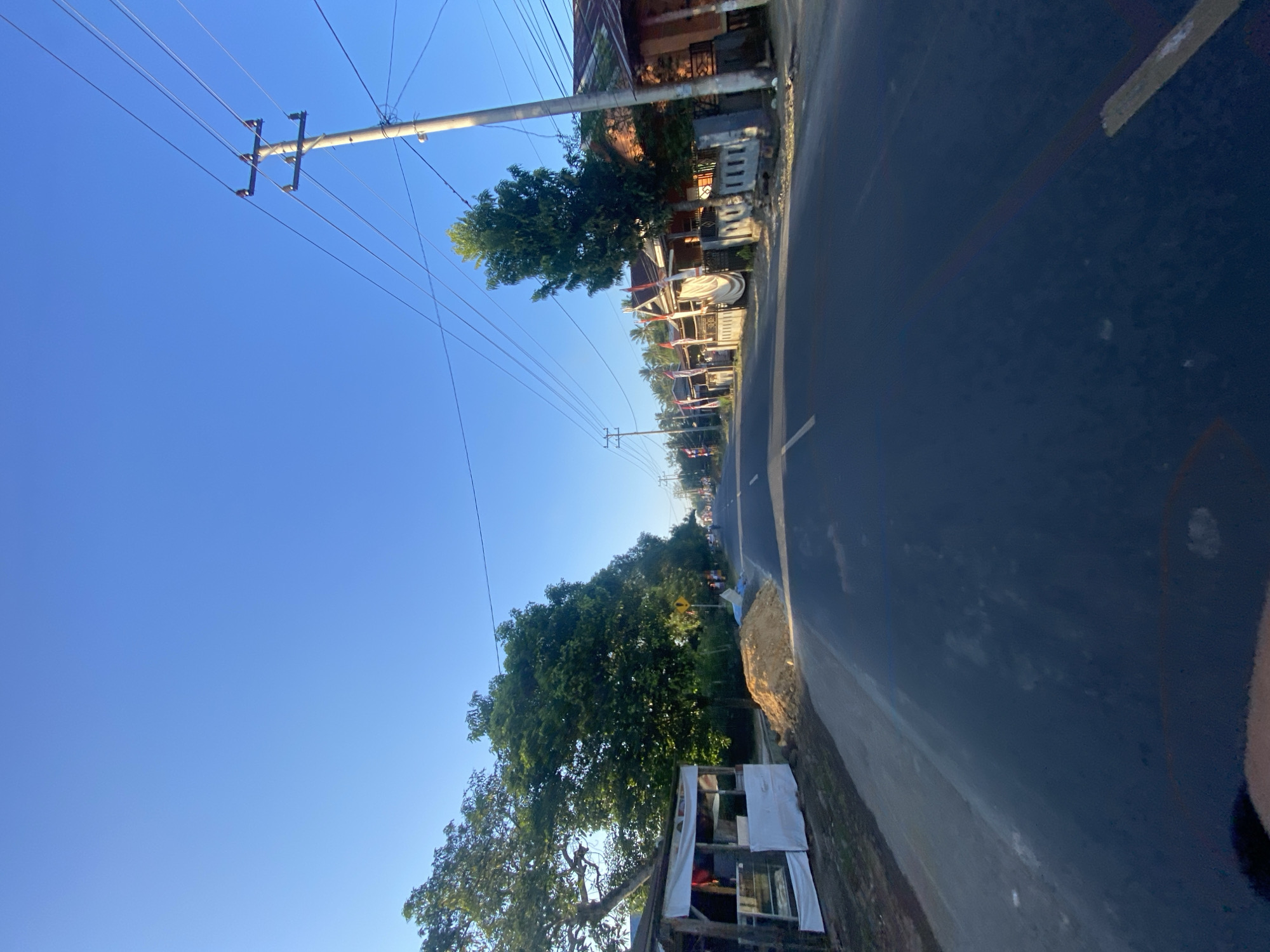 The image depicts a road in Tanah Harapan, Indonesia, under a clear blue sky. On the left side of the road, there is a small roadside stall with white tarps and a display counter. Adjacent to the stall, a pile of sand is visible. Further down the road, trees line the left side, and on the right, houses with red-tiled roofs and fences are situated, some adorned with Indonesian flags. Electrical poles with wires run along the side of the road. The road itself has white lane markings and appears to be an asphalt surface. In the distance, the road stretches towards a slightly hazy horizon, with more trees and structures visible. The scene suggests a typical rural or semi-urban setting in Indonesia during daylight hours.