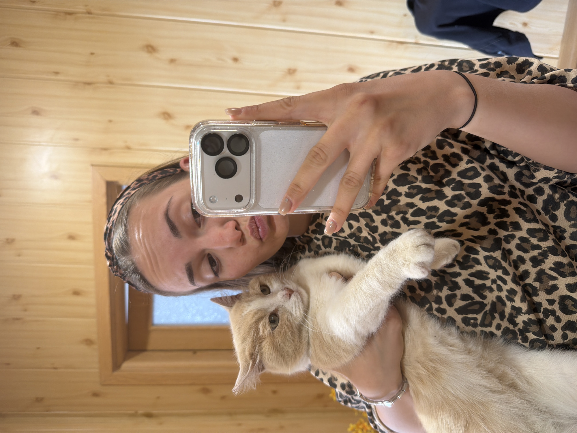 A light-skinned woman with a leopard-print headband takes a selfie with a white and tan cat. She holds a silver smartphone with a clear case in her right hand. She is wearing a leopard print shirt. The cat is being held in the woman's arms. The background consists of light brown wooden walls and a window. There is no evidence to suggest the photo was taken in Buhovo, Bulgaria.