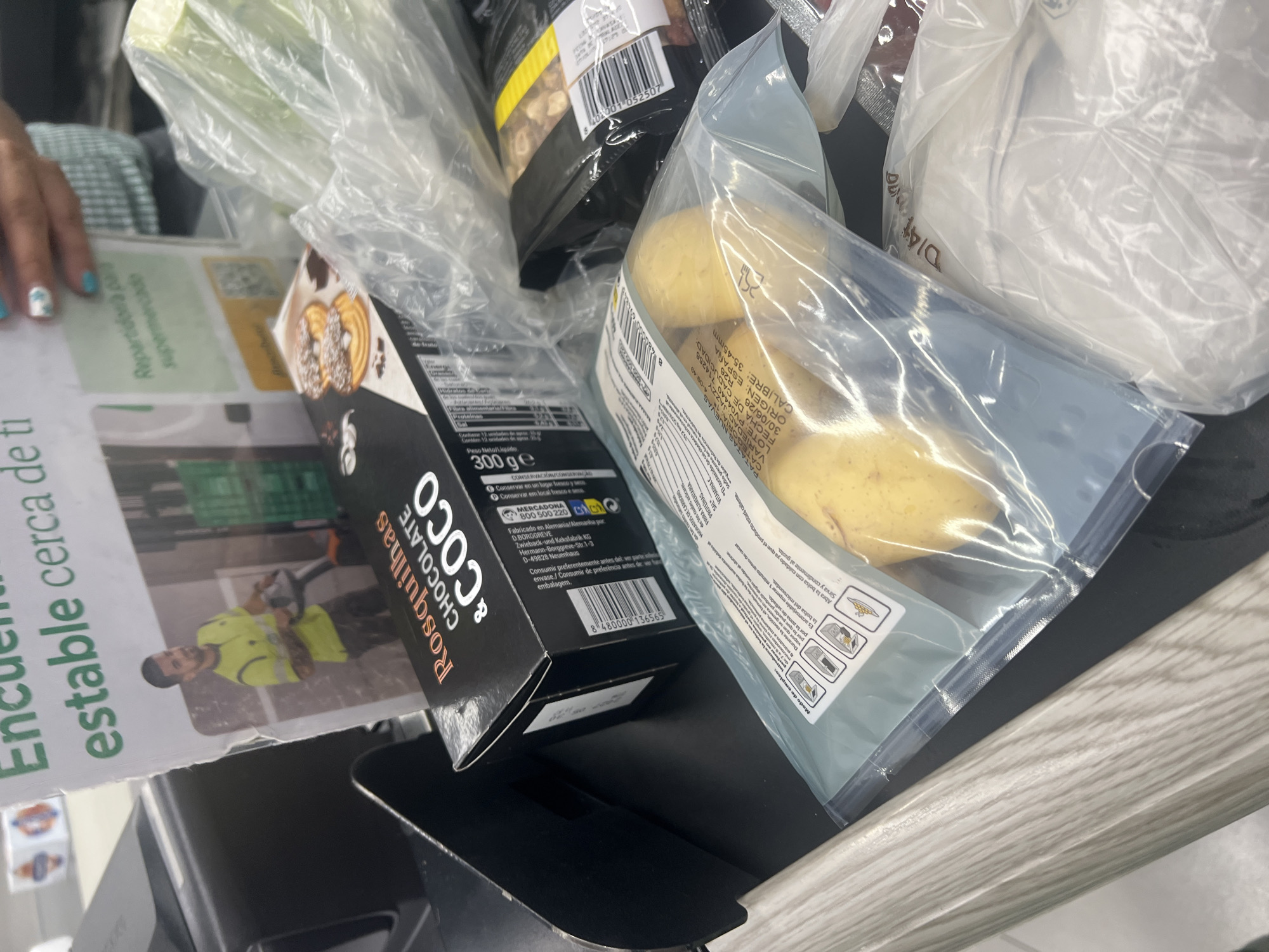 The image shows a close-up, high-angle view of groceries on a checkout counter, likely in a supermarket. The items include a box of "Rosquillas CHOCOLATE & COCO" cookies, a bag of potatoes, and other items in plastic packaging, some of which are blurry. A person's hand with blue-painted fingernails is visible on the left side of the frame, suggesting they are reaching for or interacting with the items.
The "Rosquillas" box is black with white lettering and displays an image of cookies. Text on the box indicates it weighs 300g and is "Fabricado en Alemania/Alemanha por Zwieback-und Keksfabrik KG". A barcode is also visible. The potatoes are in a clear plastic bag with text in Spanish and Portuguese, including "PATATAS NUEVAS VARIADAD JAZZY" and "ORIGEN: ESPAÑA".
A partially visible flyer or newspaper on the left has green text that reads "En", "estable cerca de ti", and "Repartidonia pala supermcate". Below this, a picture shows a man in a high-visibility yellow vest working with equipment.
The overall scene is one of everyday shopping, with the focus on the products. The lighting suggests it's daytime, and the presence of groceries and a checkout counter implies a commercial setting like a supermarket. The image appears to be taken indoors.