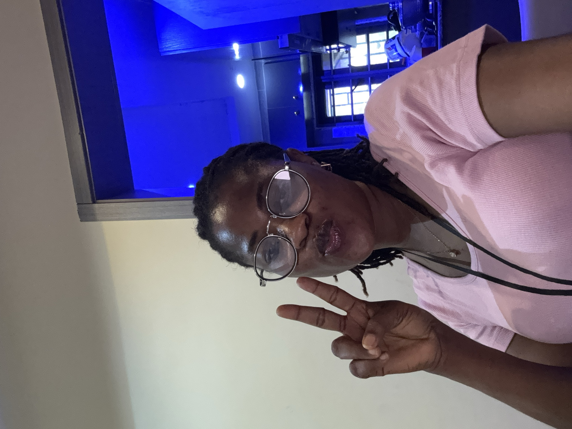 The media file is a portrait of a young Black woman wearing glasses and a pink top. She is making a peace sign with her right hand. The background is a mix of a brightly lit blue area with what appears to be shelving and light fixtures, and a plain cream-colored wall. The lighting suggests an indoor environment, possibly a bar or lounge, with the blue lights creating a vibrant atmosphere. The woman's expression is friendly and she appears to be posing for the photo. The image is oriented sideways. The provided location context is Ajara, Nigeria.