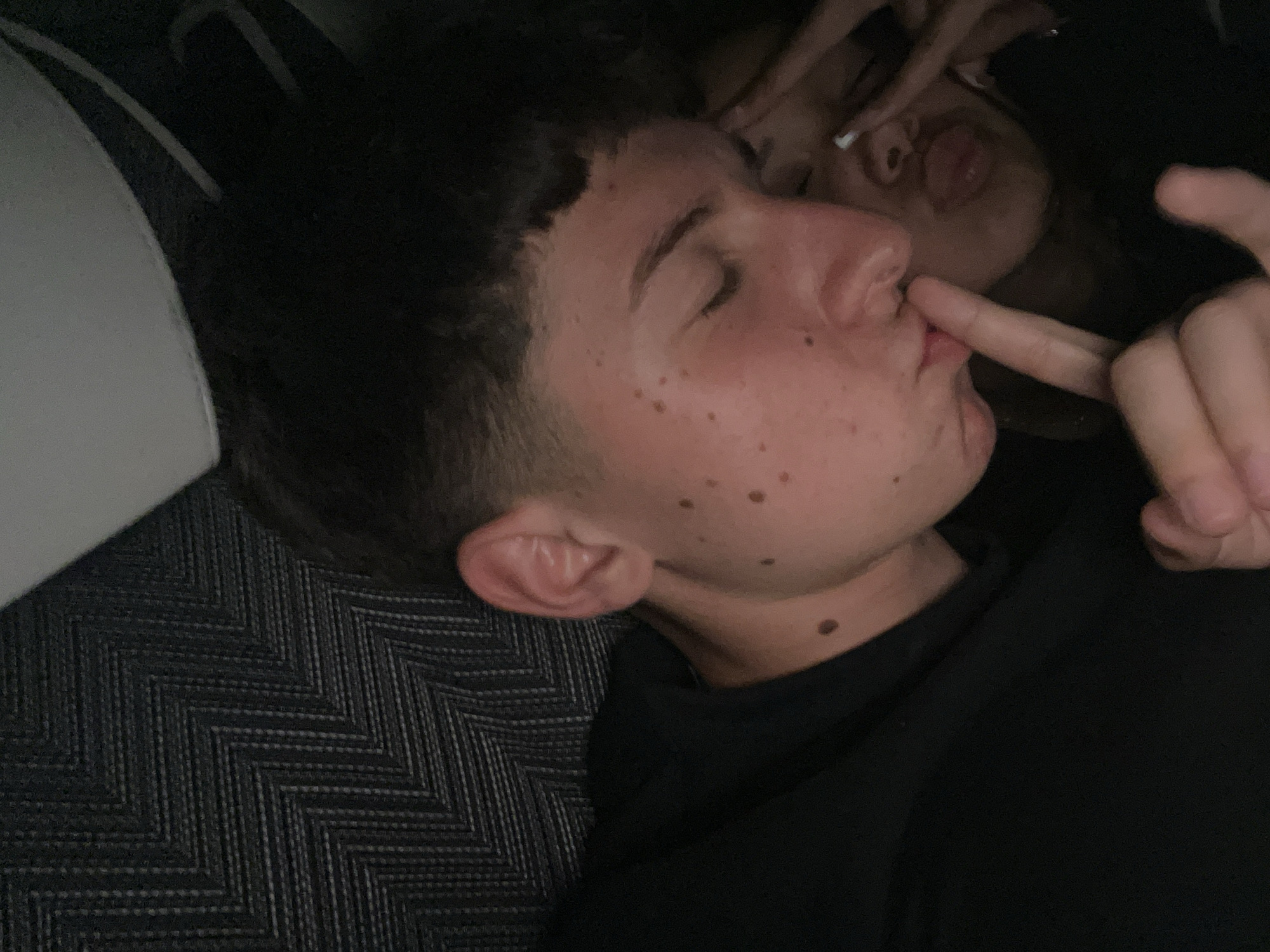 A close-up photograph shows two individuals lying down indoors on a chevron patterned fabric. The primary subject, on the bottom, is light-skinned with short dark hair, several moles on their face, and their eyes are closed. Their index finger touches their lips in a gesture. Above them, a second individual is partially visible; only their mouth and nose are clear. The background is dark and undefined. No location information can be inferred from the image.