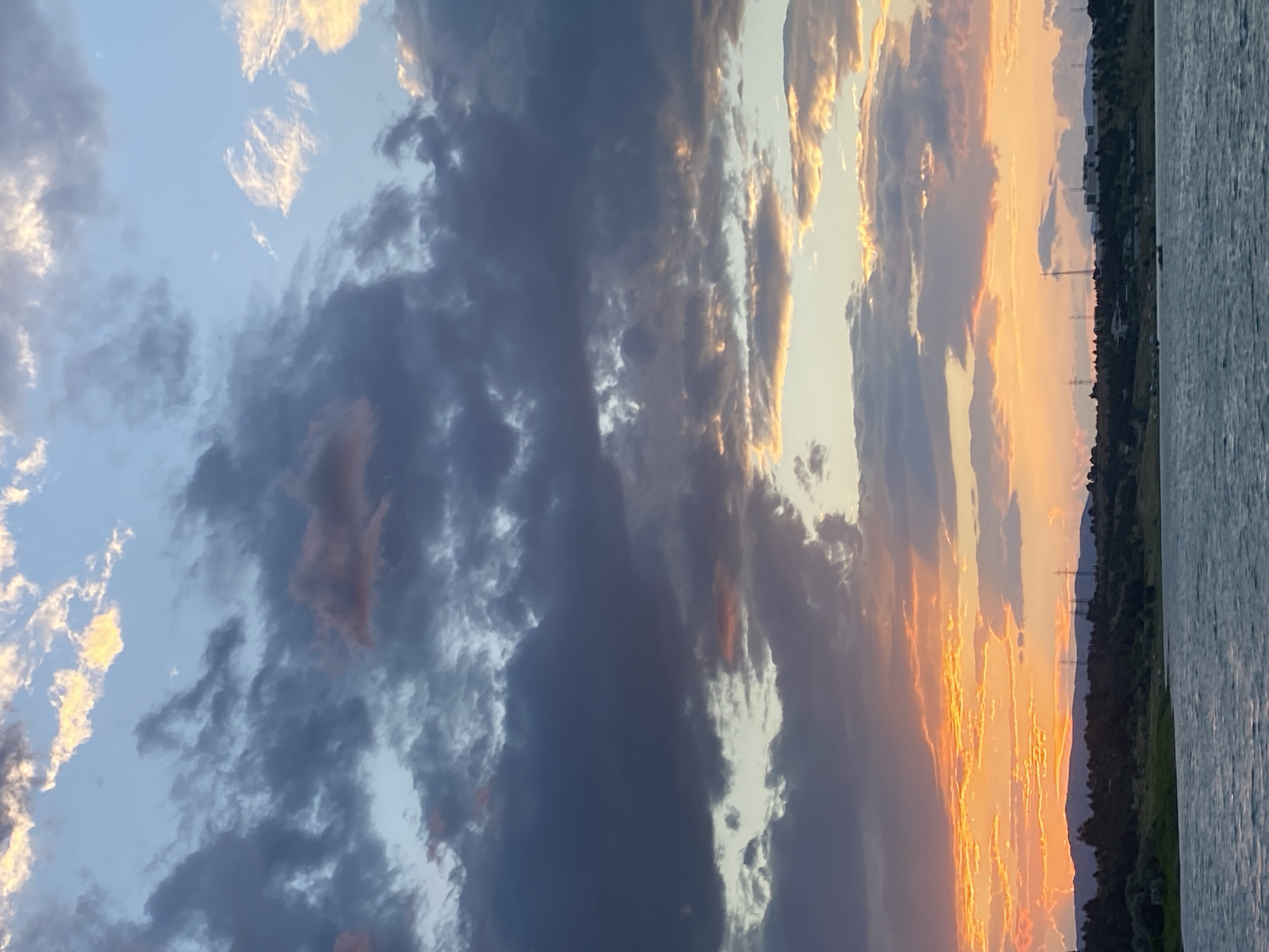 The image captures a serene sunset over a body of water, likely a lake or reservoir. The sky is a dramatic display of colors, with vibrant oranges and yellows from the setting sun illuminating the undersides of dark, cloudy formations. These clouds transition to lighter grays and blues higher up, with some wisps of white clouds catching the last rays of light.
In the foreground, the water appears rippled, suggesting a gentle breeze. The middle ground reveals a tree-lined shore with undulating hills in the distance. On the hills, several structures are visible, including what appear to be power lines or transmission towers, and some buildings that suggest a suburban or semi-urban environment.
While the exact location is not explicitly stated, the description of power lines and buildings on hills near a body of water could be consistent with areas around Tbilisi, Georgia, known for its diverse landscapes. However, without more specific landmarks, pinpointing the precise location within Tbilisi is not possible based solely on the image. No people or animals are visible in the scene, and no actions or interactions are depicted. The overall impression is one of natural beauty and quietude as day transitions to night.