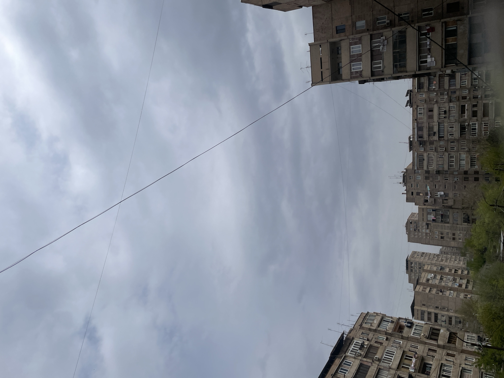 The image is taken looking up between apartment buildings in Yerevan, Armenia. The buildings are a dull, light gray color with numerous windows. There are several power lines visible crossing the sky. The sky is overcast with gray clouds. Green tree foliage is visible at the right of the frame, suggesting a nearby park or street lined with trees. Clothes are being dried on clotheslines from the balconies of some of the buildings.