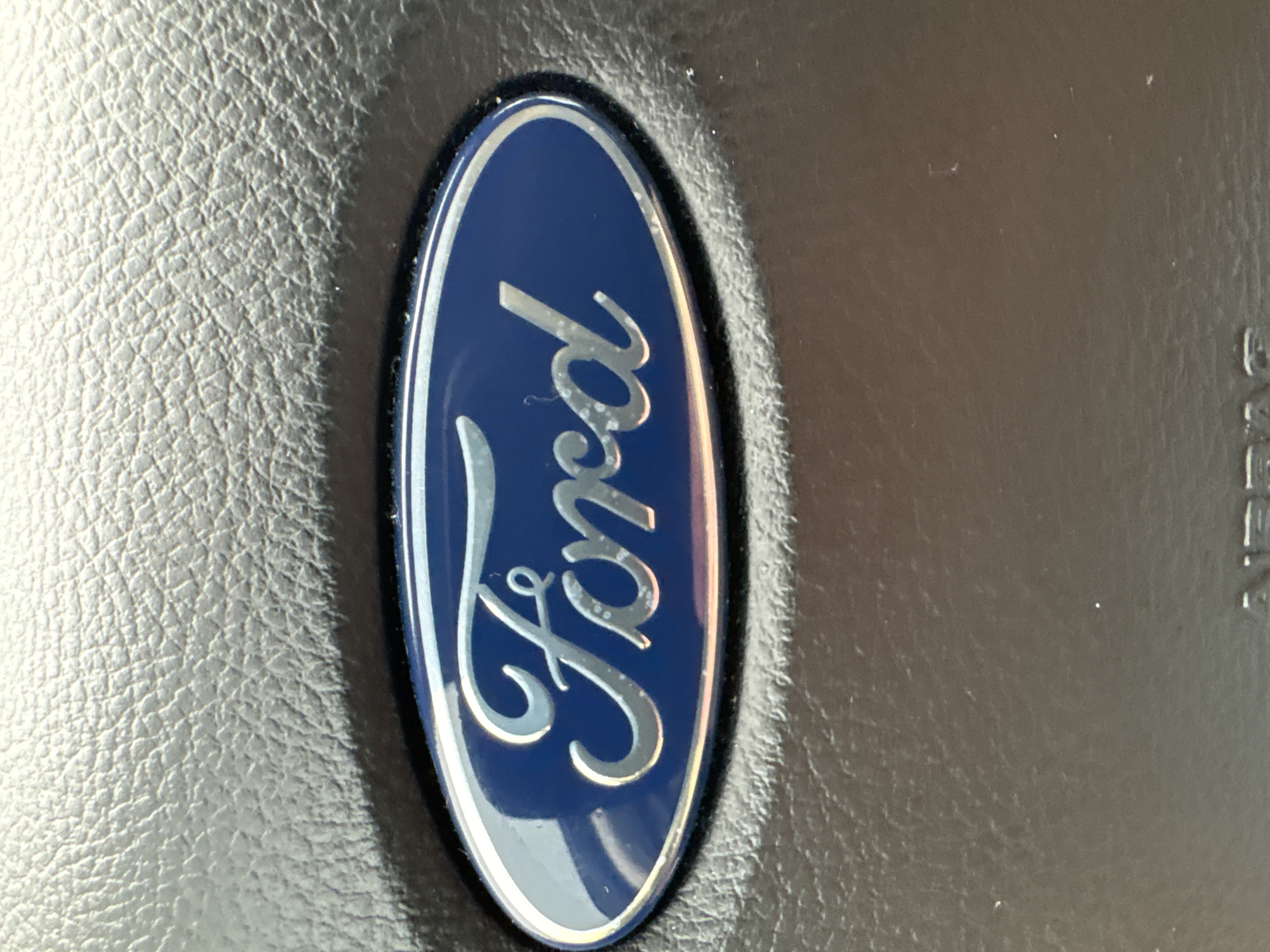 A close-up, eye-level shot focuses on the Ford logo embossed on a black, textured surface, likely a car's steering wheel or dashboard. The oval-shaped logo is a deep blue with the word "Ford" written in a silver, cursive font. The texture of the black material is clearly visible around the logo, showing small, irregular bumps. The lighting creates a slight sheen on the logo and highlights the texture of the surrounding material. There are no people or other objects visible in the frame. The image does not contain information about its location in Vitsebsk, Belarus.
