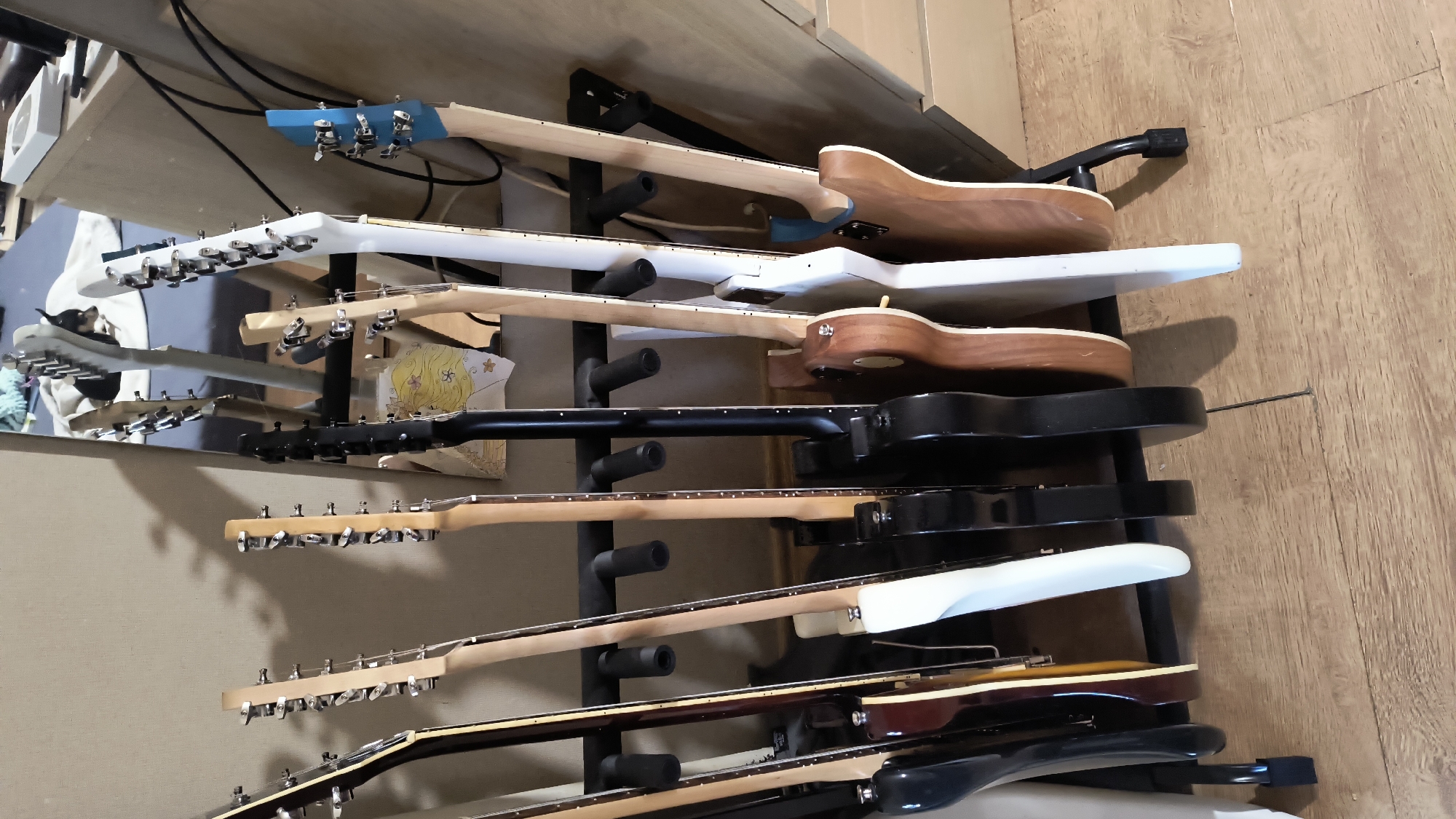 The image shows a collection of electric guitars stored vertically in a black metal guitar rack against a wall. The wall behind the rack is a light beige color. To the right of the guitar rack, there is a light brown wooden floor. A wooden cabinet with some cables on top is partially visible in the upper left corner of the image. A mirror is visible in the upper left, showing an angle of the guitars and rack with a small dog visible. The location is not specified, there is no indication that the location is in the city of Sofia, Bulgaria.