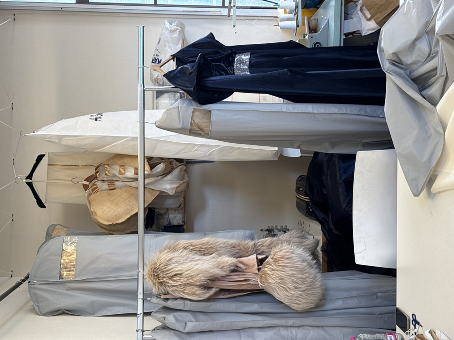 This image captures an indoor workshop or atelier in Arcueil, France, dedicated to garment production and storage. The space features light-colored walls and a window on the right providing ambient light. Two horizontal metal clothes rails are prominently displayed. The upper rail supports a grey garment bag with a clear window, a white garment bag partially marked with "A.B.", and a large, straw-colored circular textile, possibly a wide-brimmed hat, alongside other draped fabrics.
The lower, chrome-finish rail holds several additional grey garment bags. A voluminous, light brown/tan furry coat with a matching furry accessory hangs on the left side of this rail. To the right, a dark, possibly black, long garment resembling a costume or formal wear is displayed on a wooden hanger, characterized by a hooded design and a shimmering silver sequined band around the waist.
In the background, a light-colored shelf stores various items, and an industrial light blue sewing machine is visible on a table to the right, accompanied by several white thread spools. The foreground features a large white work table with a white chair pulled up to it. On the table's left, a partially consumed plastic water bottle, a dark smartphone, blue scissors, and a measuring tape are arranged. The right side of the table is covered with draped pieces of light grey and white fabric, indicating ongoing textile work. No individuals are present in the scene.