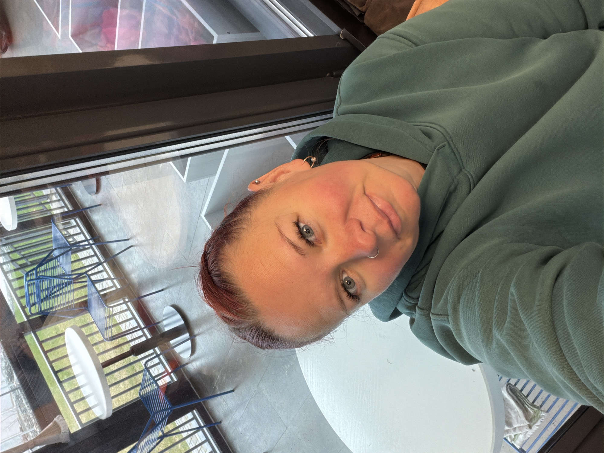 A woman with auburn hair, blue eyes, and a nose ring is taking a selfie. She is wearing a green hooded sweatshirt. The background shows a window with a view of blue chairs and round white tables. The location appears to be a public space, possibly a cafe, in Chaikovsky, Russia.