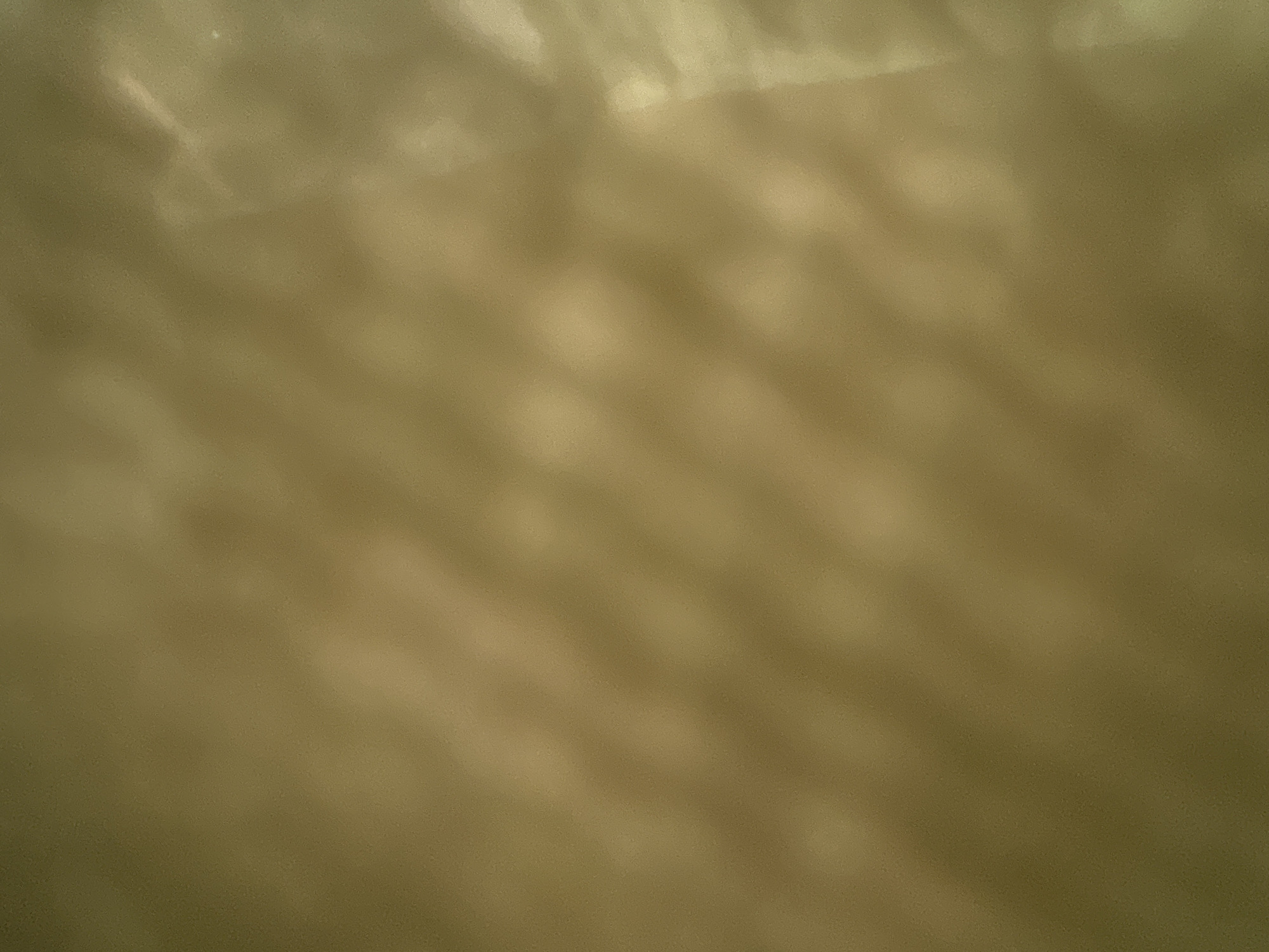 The image is a close-up, out-of-focus shot of a textured surface with a light and shadow pattern. The dominant colors are shades of brown and yellow, with some areas appearing lighter and brighter due to reflections or light sources. The texture appears to be a grid-like pattern or a repeating arrangement of raised or indented elements, casting shadows that create a dappled effect across the surface. The lighting creates highlights and darker areas, suggesting a rough or uneven material. There are no people, specific objects, or discernible environmental features that can be identified. The location in the city of Kalitsino, Russia, cannot be determined from the visual information provided in the image.