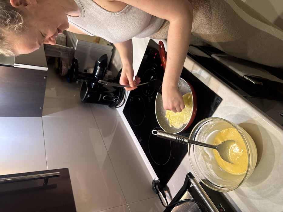 A woman is cooking in a kitchen. She is making crepes in a red pan on a black stovetop. Beside the stovetop is a bowl with more batter and a spoon. A food processor is nearby on the counter. The kitchen has light-colored tile flooring and sleek cabinetry.