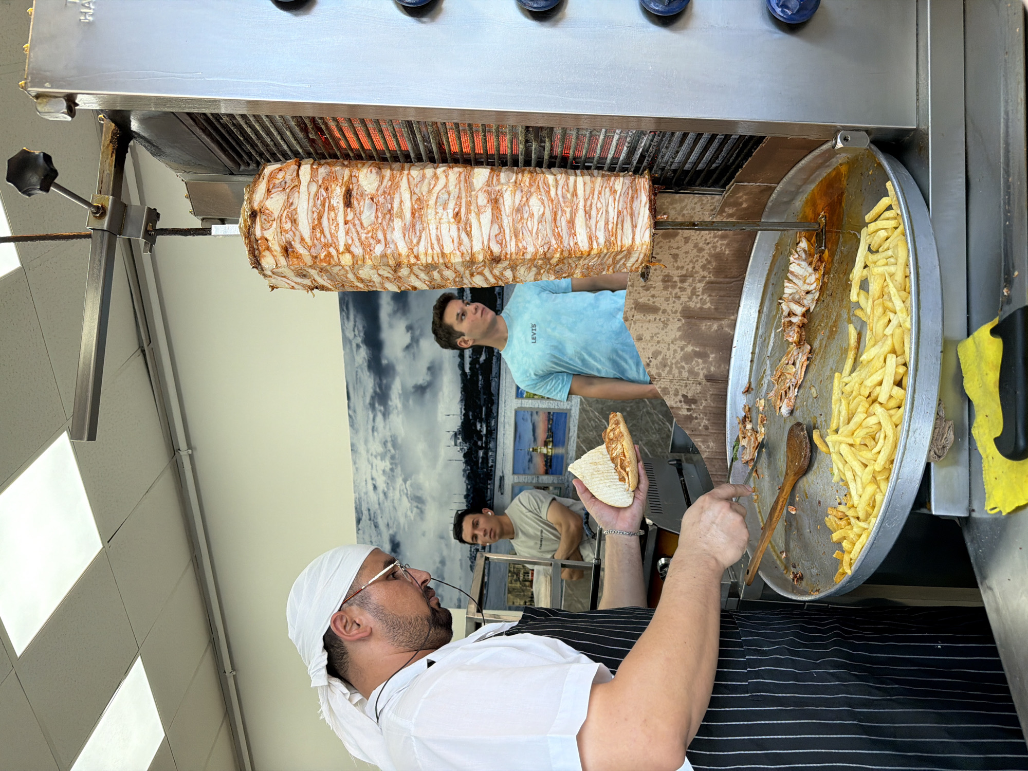 In Kadıköy, Turkey, a cook wearing a white headscarf and apron prepares a doner kebab. He is holding a half-eaten pita bread in his left hand, which he is about to fill with meat sliced from a rotating vertical spit. The spit, laden with seasoned meat, is heated by glowing red coils. Below the spit, a large metal tray holds a generous portion of french fries and shaved doner meat. In the background, two young men are visible. One, in a light blue t-shirt, stands to the right of the doner spit, looking towards it. Another man, in a grey t-shirt, stands further back, his arms crossed. A mural depicting a cloudy sky and city skyline adorns the wall behind them. The setting appears to be a small eatery or food stall.