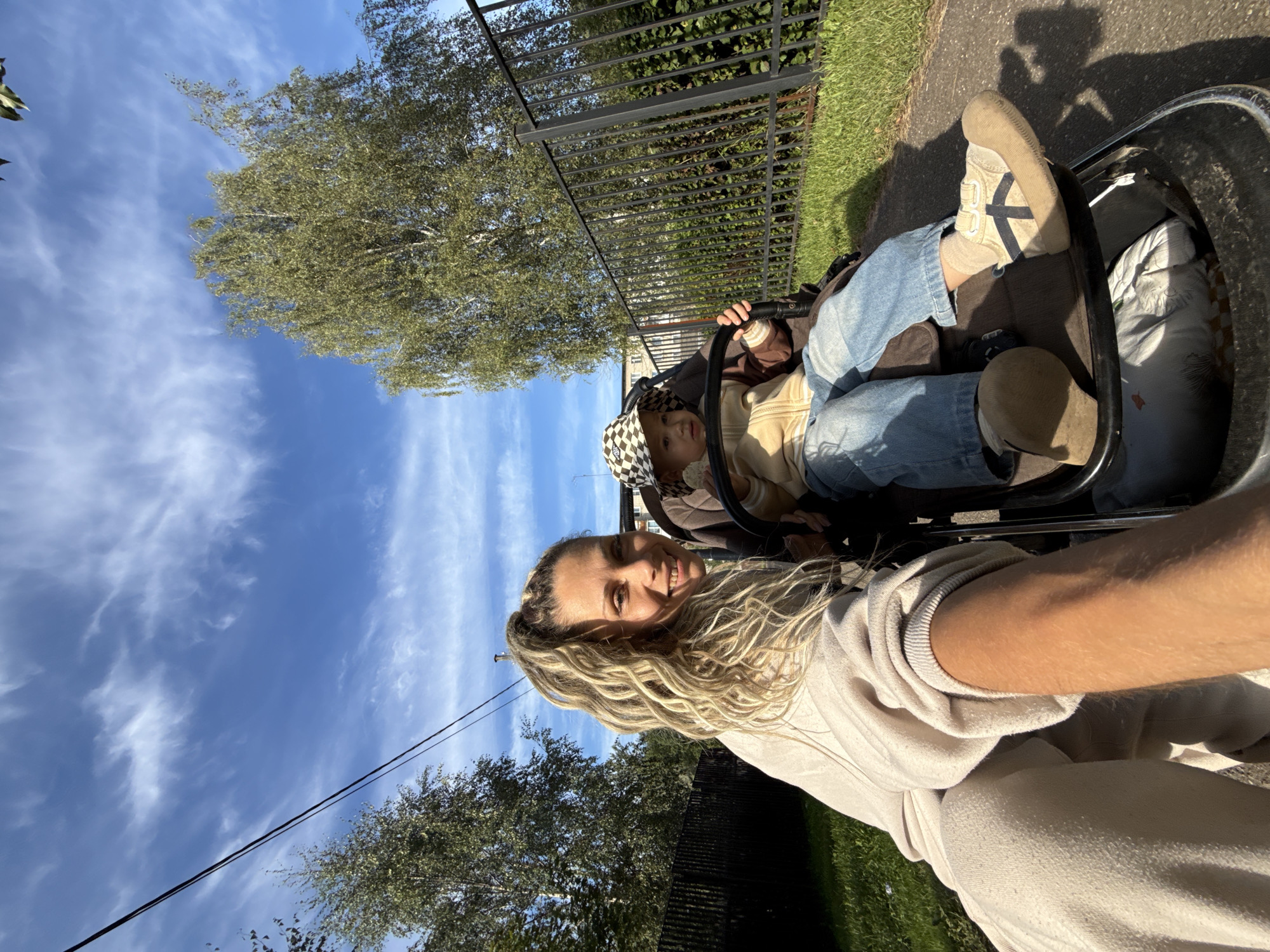 In Zhukovo, Russia, on a sunny day with wispy clouds, a woman with blonde, dreadlocked hair pulled into a ponytail is selfie-taking while sitting in a stroller. Her left arm is extended, holding the phone. She wears a cream-colored hooded sweatshirt and matching sweatpants. Beside her, a toddler in a stroller wears a checkered bucket hat, a light brown zip-up jacket over a tan t-shirt, and blue jeans. The toddler's feet are clad in light-colored sneakers with dark blue accents and beige socks. The stroller has a black frame and brown fabric. Behind them, a black metal fence runs along a grassy area with dense green foliage. A tall tree with green leaves stands to the left, and another tree with sparser foliage is visible behind the fence. Overhead, power lines stretch across the bright blue sky.