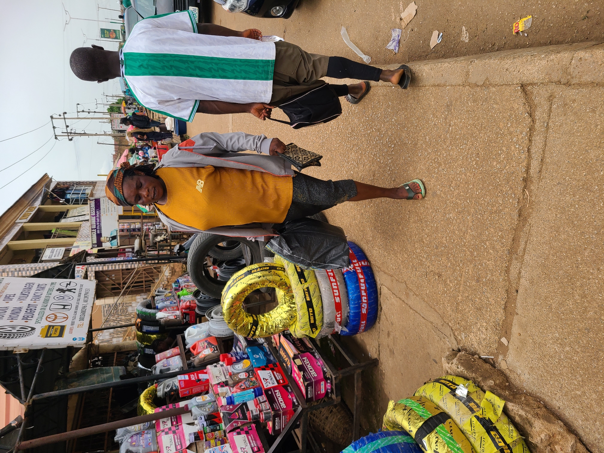The image depicts a street scene in an urban area, possibly a market area based on the presence of shops and roadside vendors. A woman with a headscarf, wearing a mustard yellow shirt and grey jacket, stands near a display of tires and auto parts. To her right, a man in a white and green striped shirt walks along the sidewalk. The area is crowded, with various items for sale, including tires wrapped in colorful plastic. There are buildings and power lines visible in the background. The location appears to be in the city of Unknown, Unknown, based on information from the OCR, and the presence of a sign for NERAT PLAZA ALONG BAIKIN JAD KOAL.