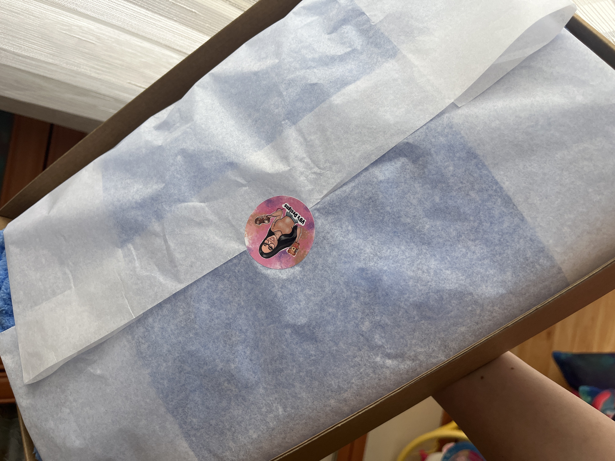 A cardboard box is open, held by a person's arm, and filled with layered white and blue tissue paper. A round sticker with a cartoon image is on the blue paper. The location appears to be indoors, based on the lighting and nearby objects. No specific location in Pernik, Bulgaria, is evident from the image.
