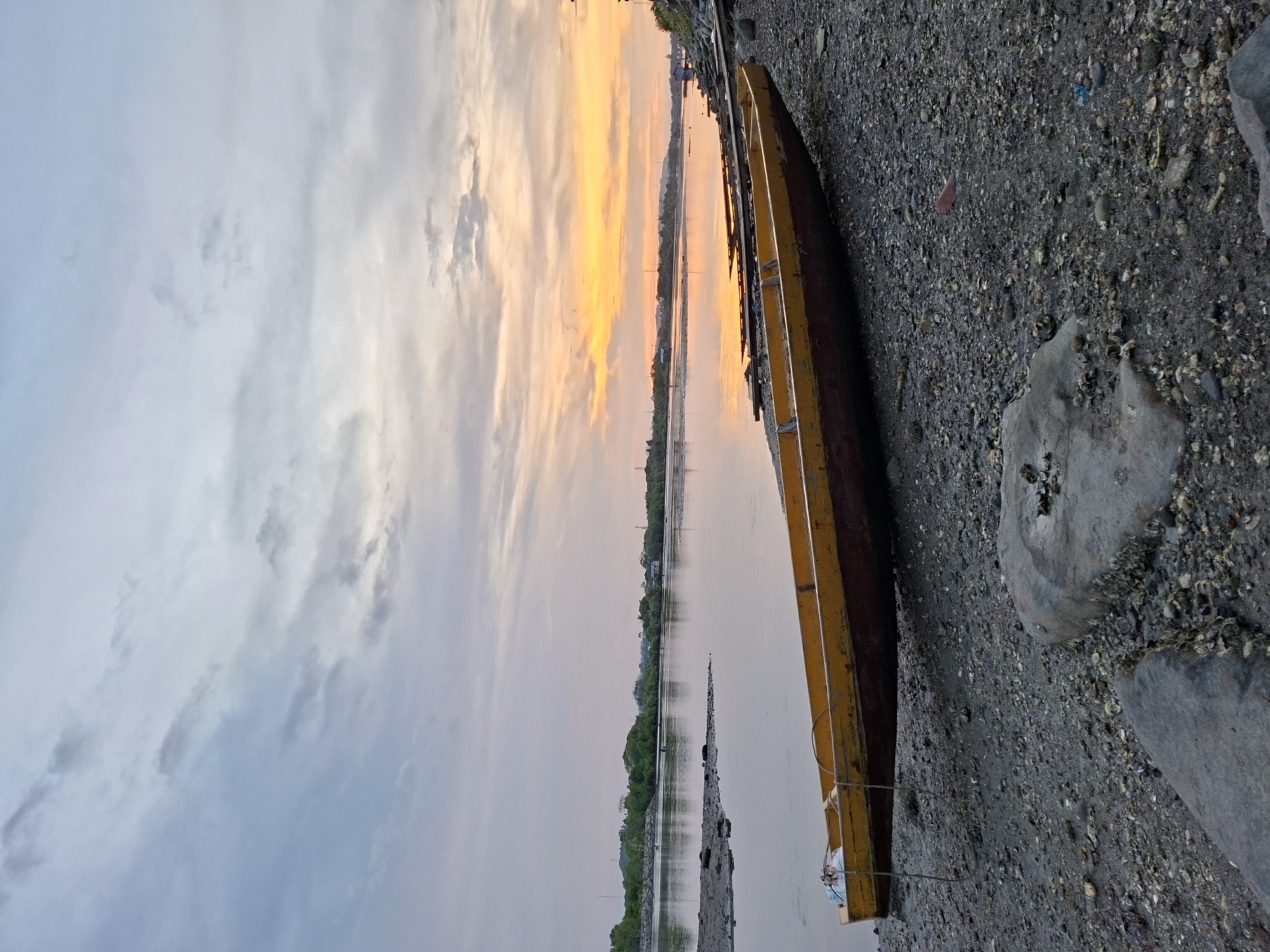 A horizontal, scenic shot shows a yellow boat aground on a gray, pebbly beach at dusk or dawn in Lhokseumawe, Indonesia. The sky is a blend of gray, white, yellow, and orange, with clouds scattered across. The water reflects these colors, creating a smooth, mirrored surface. Across the water, a green shoreline is visible. The boat rests on the right side of the frame, parallel to the horizon, with two gray rocks next to it, covered in small, dark shells. The beach stretches toward the water, a mix of small pebbles, sand, and dark debris. No people or other boats are visible.