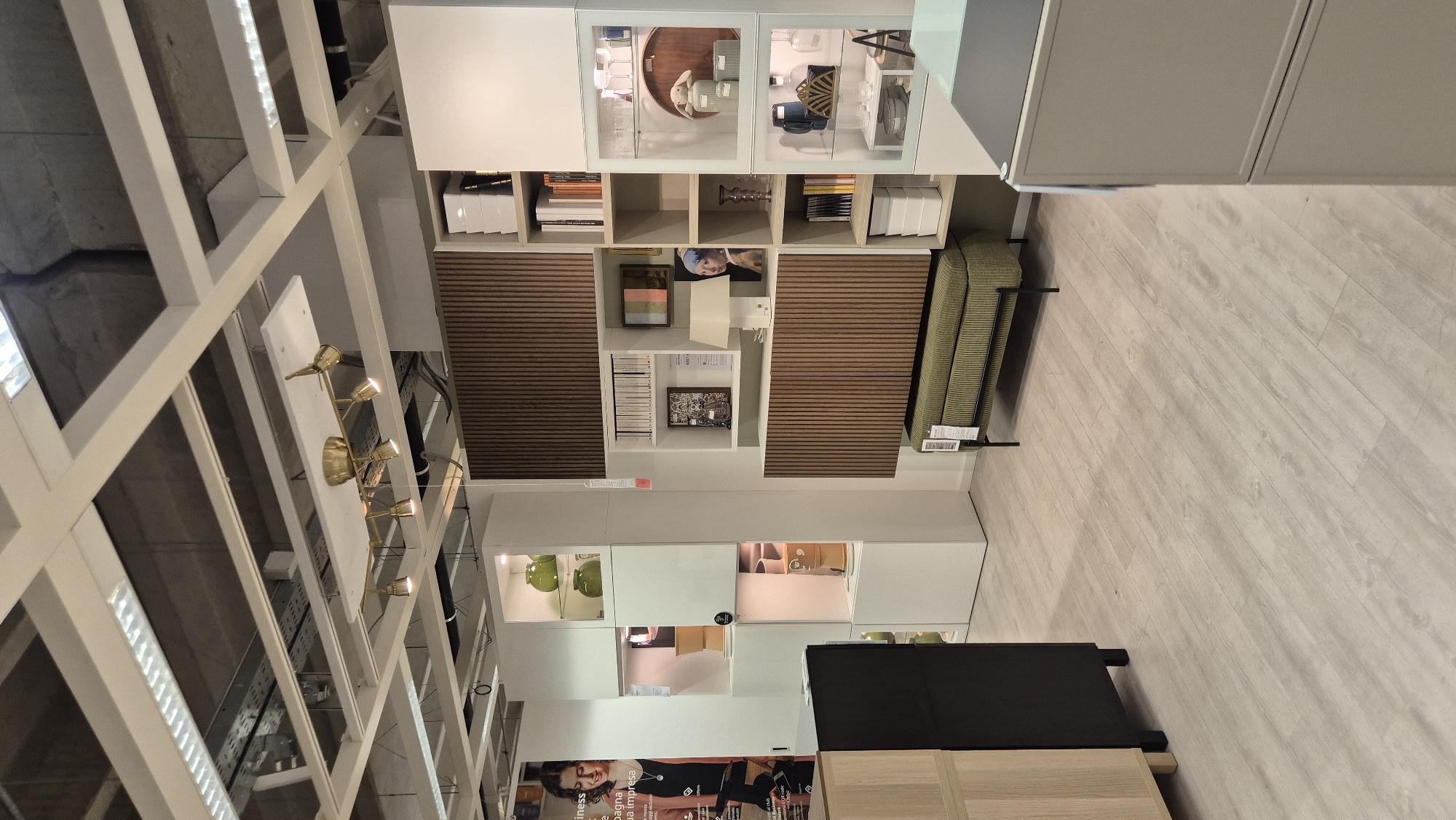The image depicts a retail showroom, likely a furniture store, with a focus on home decor and display units. The showroom features a modern aesthetic with light-colored laminate flooring and a white grid ceiling system with integrated lighting.
In the foreground, a beige cabinet with a dark countertop is partially visible on the right. To the left, a black cabinet with dark legs stands adjacent to a light wood cabinet.
The central area of the image is dominated by modular shelving units. One unit, with a dark, slatted wood facade, contains various decorative items, including books, a lamp, and a framed print that appears to be Vermeer's "Girl with a Pearl Earring." Below this unit, a green upholstered bench with black metal legs is positioned.
Another white modular shelving unit to the left displays items such as green vases, yellow decorative objects, and stacked white boxes. A poster featuring a smiling woman and text in Italian is mounted on the wall to the far left.
The overall environment suggests a well-lit and organized commercial space designed for product display and customer browsing. There are no people visible in the scene. The image does not contain any specific visual cues that would indicate the location in Baronissi, Italy.
