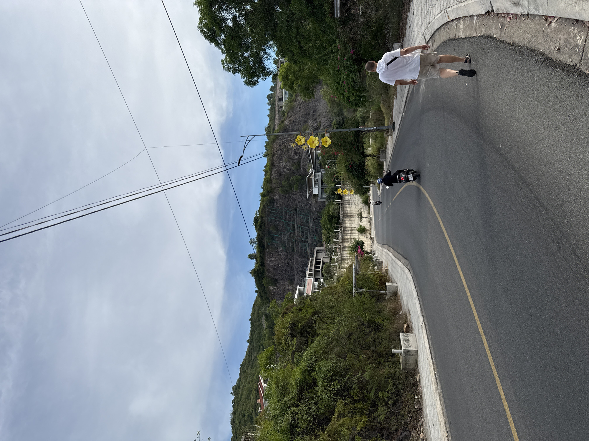 The image depicts a scenic road winding through a hilly, green landscape in Nha Trang, Vietnam. A man in a white t-shirt and beige shorts walks away from the camera on the right side of the road, while two people on motorcycles travel in the opposite direction. The road is asphalt with a yellow line marking the center. On the left, lush green vegetation covers a steep hillside dotted with buildings. In the background, a rocky cliff face is visible, with a series of green nets secured to it, likely for erosion control. Yellow flower-like decorations hang from a lamppost on the right side of the road. The sky is partly cloudy, with visible power lines crossing the frame. The overall atmosphere is one of a sunny day in a tropical environment.