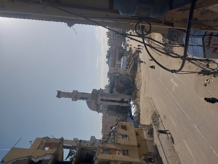 The image depicts a scene of extensive urban damage in Khan Yunis, Palestinian Territory, centered on the Great Mosque of Khan Yunis. The mosque's tall, white minaret and dome are visible in the mid-ground, though the main structure behind the minaret appears significantly damaged and partially collapsed.
Buildings on both sides of a paved street exhibit severe destruction, with multi-story structures having large sections of their facades missing, exposing interior debris and reinforcing bars. Piles of rubble, concrete fragments, and twisted metal are scattered along the road and accumulate at the base of the damaged buildings.
A two-lane paved road, marked with white lines and a right-turn arrow, traverses the scene. Its surface shows patches of dirt and debris.
Several individuals are present on the street: one person walks on the left side, another person is near the bottom center, and a group of approximately 5-7 people walks further down the street on the right. Another person on the right pushes a dark two-wheeled cart or similar object, with a smaller figure nearby. No vehicles are visible on the immediate road.
The sky is clear and bright, with a strong sun glare originating from the upper right quadrant. The photograph is taken from an elevated position, with dark cables and a large water dispenser partially visible in the immediate foreground on the right, indicating a vantage point from within a damaged building. A white box with Arabic text is also partially visible at the bottom right.