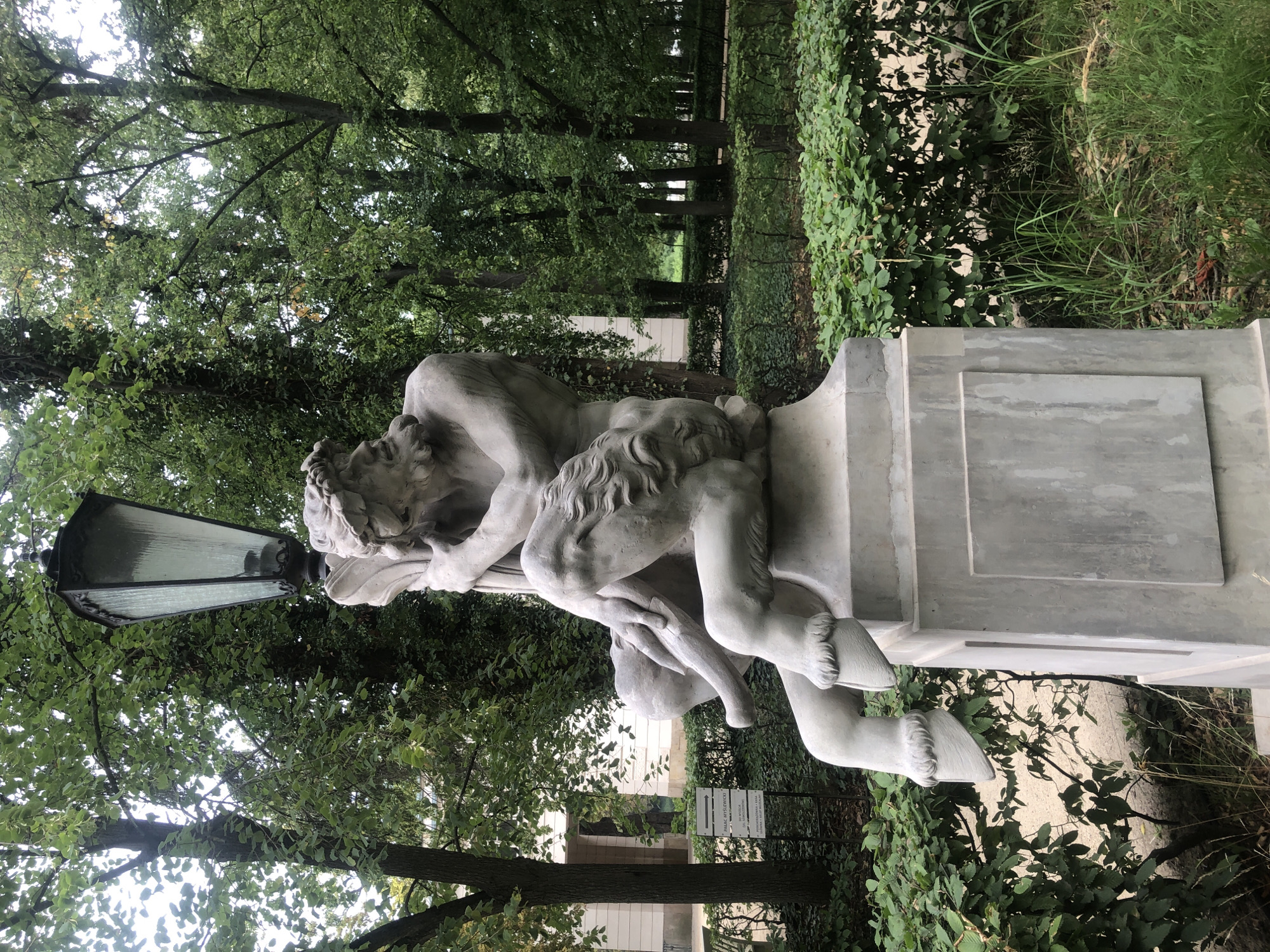 A stone sculpture of a satyr is positioned on a pedestal in the Saxon Garden in Warsaw, Poland. The satyr is depicted in a seated position, with his arms wrapped around a lamppost. He has a beard and is adorned with a wreath of leaves on his head. The sculpture's lower body features hooves, indicating his mythical nature. The background is a lush green park with trees and manicured hedges, suggesting a temperate climate. A building with white walls is visible behind the sculpture, partially obscured by greenery. A signpost with Polish text is also present to the left of the sculpture. The lighting suggests an overcast day.