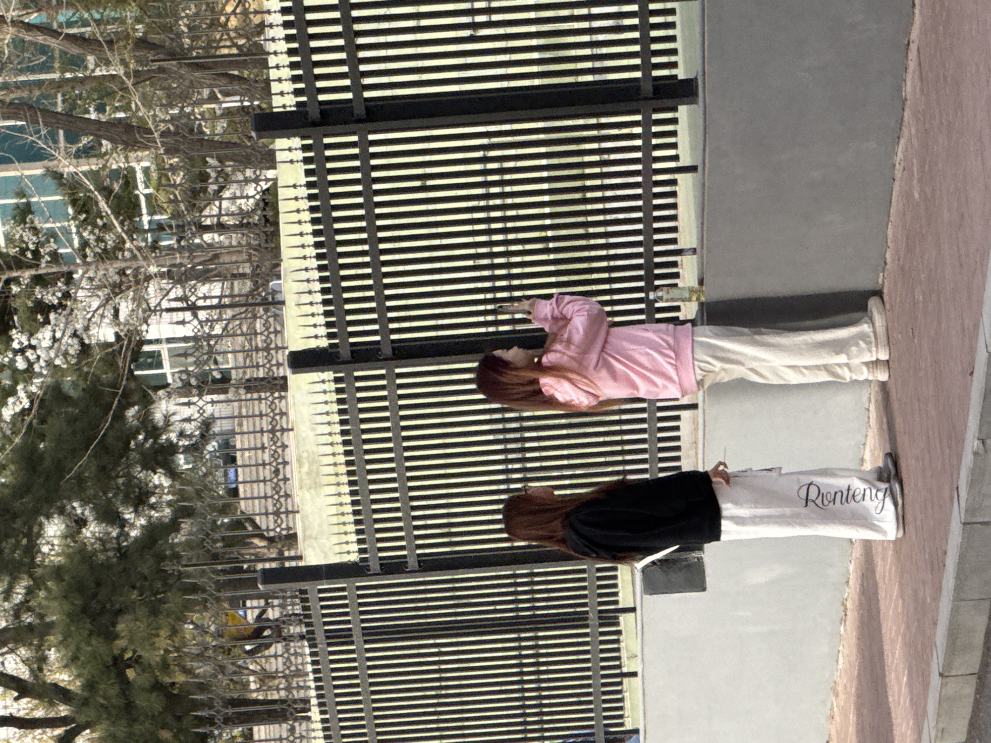 Two young women stand near a black iron fence with horizontal bars in Zhongtun, China. One woman wears a pink hooded sweatshirt and light-colored pants. She is looking at her phone. The other woman wears a black jacket and white pants with "Runteng" printed on the leg. They are standing on a sidewalk. Behind the fence, there are trees, and a building with many windows is visible in the distance.