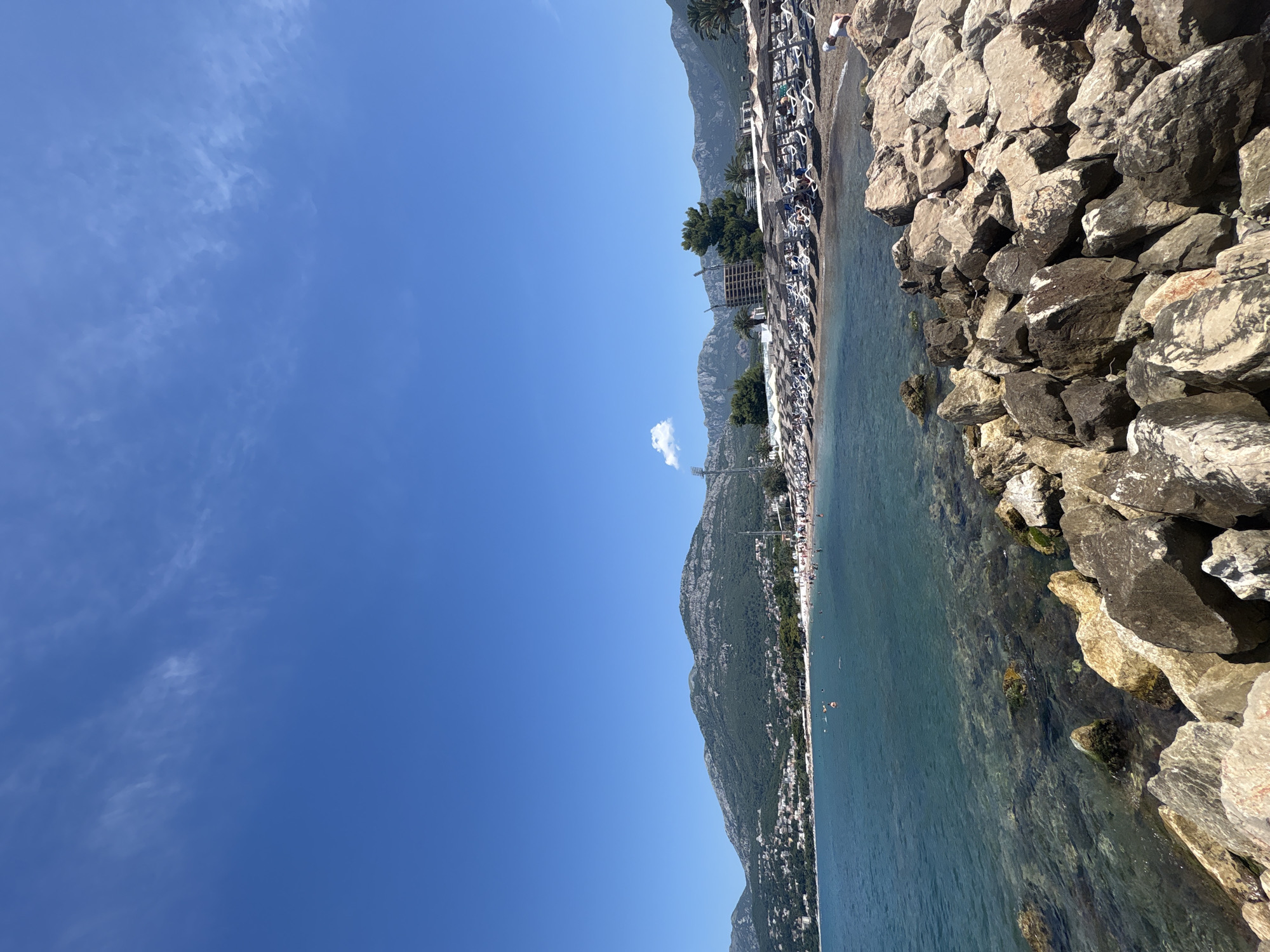 A wide-angle shot captures a sunny day at the beach in Bar, Montenegro. In the foreground, a rocky shoreline meets the clear, shallow water. Beyond the water lies a crowded beach with rows of white lounge chairs and thatched umbrellas, dotted with numerous sunbathers. The beach is backed by a line of trees and a town built into the hillside, leading up to a large, verdant mountain range that dominates the background. The sky is a vibrant blue with a few wispy clouds. There is also a construction site visible on the right side of the beach.