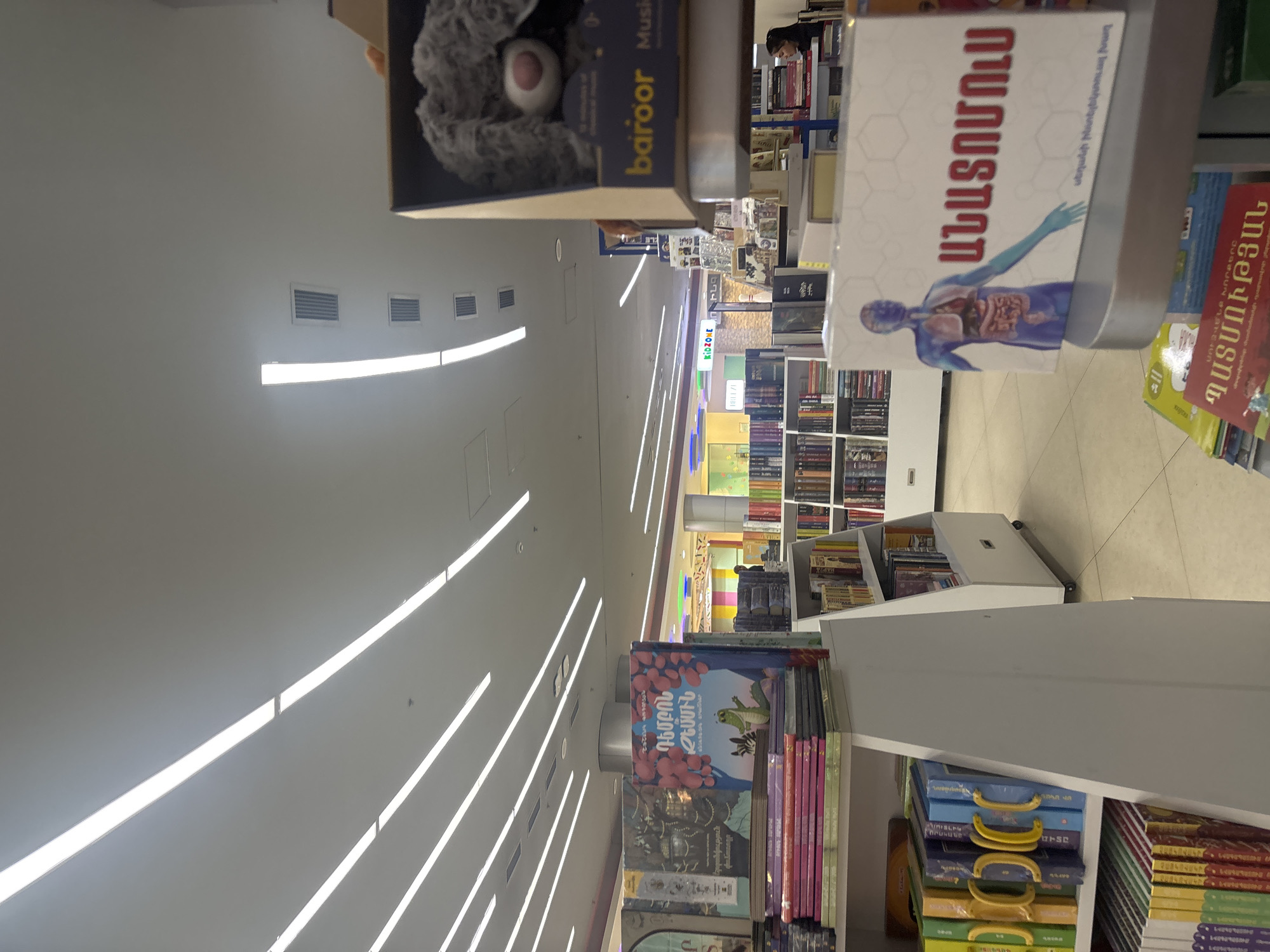This upward-angled image captures the interior of a brightly lit bookstore or a book section within a larger store, located in Yerevan, Armenia. The scene is dominated by numerous bookshelves filled with a wide variety of books, extending into the background. The ceiling is white and features several long, linear fluorescent or LED light fixtures, along with rectangular ventilation grilles, contributing to a modern and well-illuminated environment.
In the foreground, books are prominently displayed. One notable book has a white cover with an illustration of the human body, revealing internal organs, and the Armenian title "ԱՆԱՏՈՄՈւՍ" (ANATOMUS). Nearby, another book has "ՊԱՏՄՈՒԹՅԱՆ" (PATMUTYAN - History) visible on its spine, and text indicating it's an "սեղանի ինտելեկտուալ խաղ" (table intellectual game). A book with a grey teddy bear on its cover also features the word "baroor."
Further into the store, a children's book with a vibrant illustration of a frog and the Armenian title "ԴԵՄԲՈՆ" (DEMBON) is visible on a shelf. A "KIDZONE" sign indicates a dedicated area for children's literature. Many other books with colorful spines and various sizes line the shelves. A person's head is partially visible in the background, suggesting the presence of shoppers or staff. The abundance of books with Armenian text confirms the location context of Yerevan, Armenia.