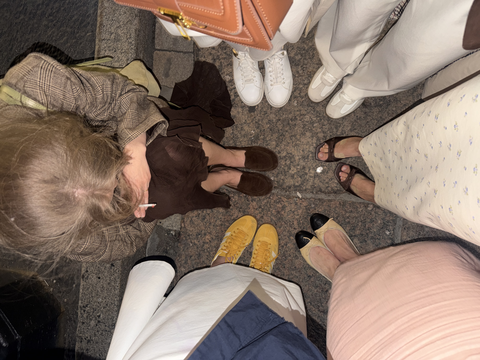 A high-angle medium shot captures a group of five individuals, viewed from the waist down, standing on a cobblestone street in Saint Petersburg, Russia. The individuals are arranged in a circle, their legs and footwear the primary focus. In the foreground, a person wearing a white skirt and dark blue shorts stands with bright yellow Adidas sneakers. To their right, another individual is adorned in a pale pink skirt and beige ballet flats with black cap toes. Moving clockwise, the next person is wearing a white floral dress and dark brown square-toed sandals. Further around, a person in light gray trousers wears white and gray low-top sneakers, while another individual in similar light gray trousers sports white lace-up sneakers. Finally, to the left, a person in a brown plaid jacket and dark brown skirt is partially visible, their legs crossed, wearing brown suede loafers. A lit cigarette is held in one person's hand, near their face, which is also visible in the upper left quadrant of the image. A brown leather handbag rests at the top of the frame. The ground appears wet, reflecting the ambient light.