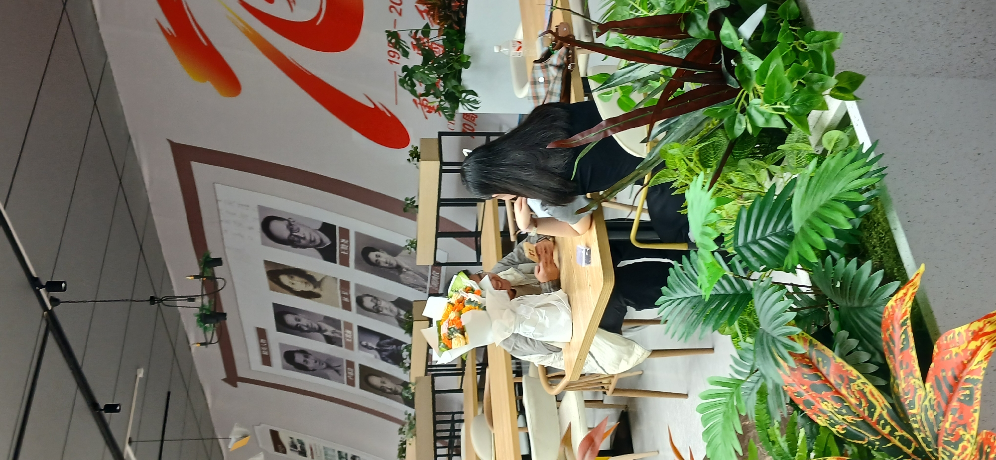 In the Chongqing High-tech Zone, China, two individuals are seated at a wooden table in what appears to be a restaurant or cafe. A bouquet of flowers, wrapped in white paper, is placed on the table. One person, on the left, is partially obscured by the bouquet and is looking down at their phone, smiling. The other person, on the right, has long dark hair and is facing away from the camera, also looking down. The environment features a wall decorated with framed portraits of several individuals in black and white. A large banner with red and orange abstract shapes and text that includes "1959 - 20" is also visible on the wall. Potted plants, including large, tropical-looking foliage, are arranged in planters in the foreground and on the wall. The lighting appears to be artificial, likely from overhead fixtures.