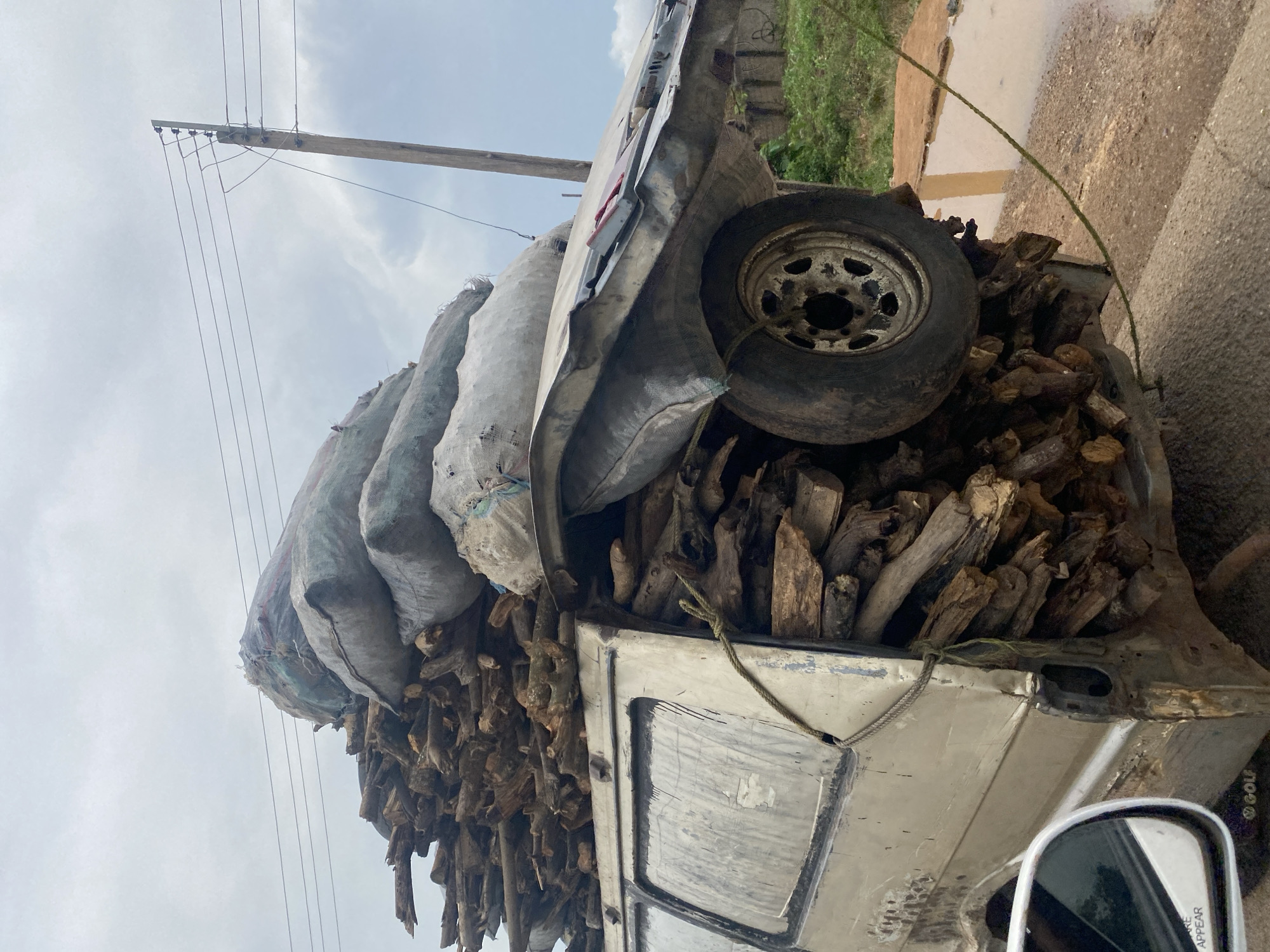 The image shows a severely overloaded and damaged van in Karshi, Nigeria. The van, a light-colored minivan, is packed to overflowing with firewood and large sacks, presumably containing agricultural produce or other goods. The firewood is stacked precariously high on the roof and sides of the vehicle, and the sacks are piled on top of the wood. The rear passenger door of the van is open and filled with firewood. The roof of the van is significantly damaged and appears to be partially collapsed, with a spare tire wedged on top, contributing to the overall instability of the load. A rope is tied around the van, attempting to secure the cargo. The scene is set against a backdrop of a cloudy sky, a wooden utility pole with multiple wires, and some vegetation. The ground appears wet and muddy in places, suggesting recent rain. A side mirror of another vehicle is visible in the lower left corner of the frame, indicating the perspective is from another car on the road. The overall impression is one of extreme overloading and disrepair of the vehicle, posing a significant safety hazard.
