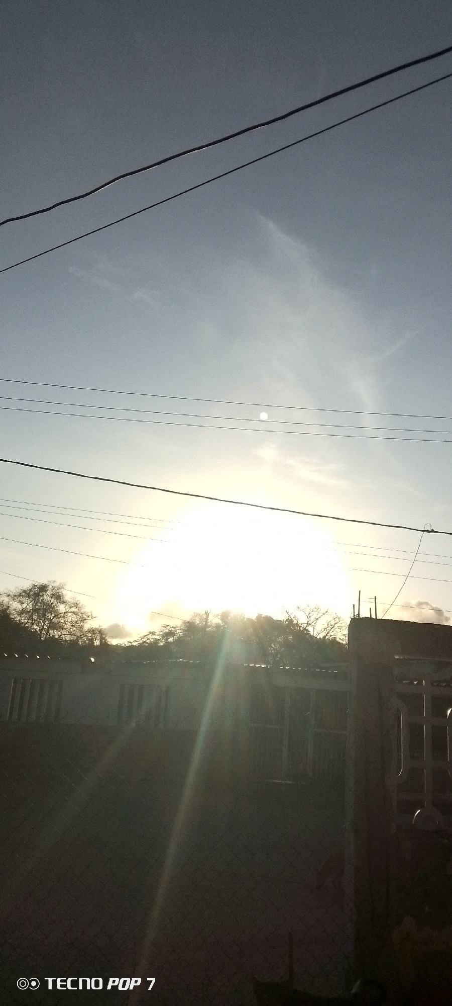 The image shows a bright sunrise or sunset, possibly taken in Altagracia, Venezuela. In the foreground is a building with a fence, obscured in shadow due to the bright light source behind it. The light appears to be coming over a line of trees or a treeline. Overhead, several power lines run across the sky, and some clouds are visible surrounding the bright light source, which could be the sun. The bottom left corner displays the watermark "TECNO POP 7," suggesting the image was taken with that specific phone model.