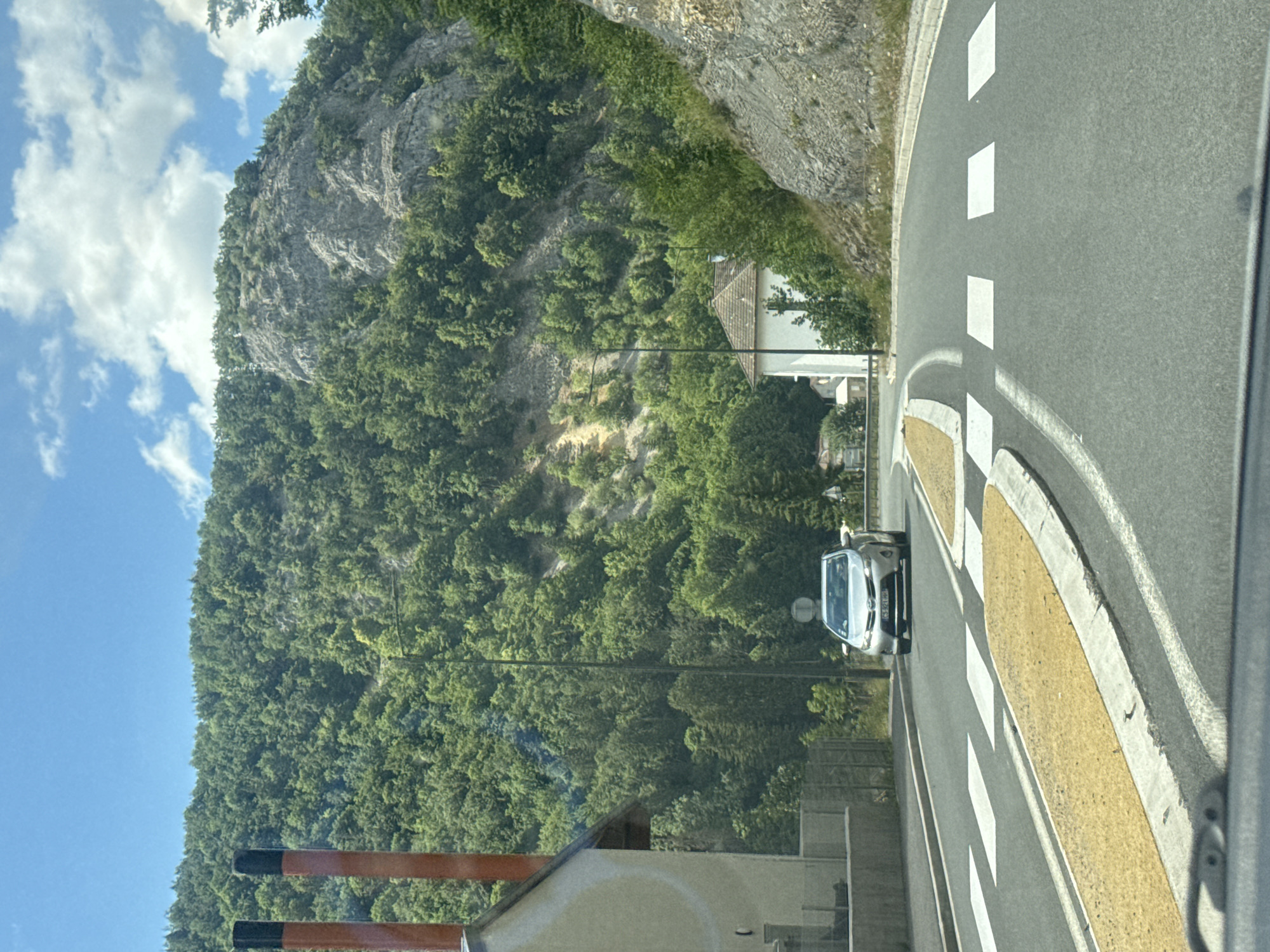 The image captures a view from inside a vehicle, looking forward through the windshield at a paved road. The road curves to the right and features a pedestrian crossing with white stripes and yellow painted islands. A silver car is visible on the road, approaching the pedestrian crossing. The surrounding environment is mountainous and lush, with dense green trees covering the slopes of a large hill that dominates the background. A rocky cliff face is also visible on the right side of the hill. On the left side of the road, there is a light-colored building with a dark-roofed section and two prominent reddish-orange chimneys with black tops. Further in the distance, another white building with a tiled roof can be seen nestled amongst the trees on the mountainside. The sky is blue with scattered white clouds. The image appears to be taken on a clear, sunny day. The scene is consistent with a road passing through a scenic, possibly rural or suburban area in a mountainous region. Given the context of Hauts de Bienne, France, this location is likely a road leading into or out of the town, showcasing its natural surroundings.
