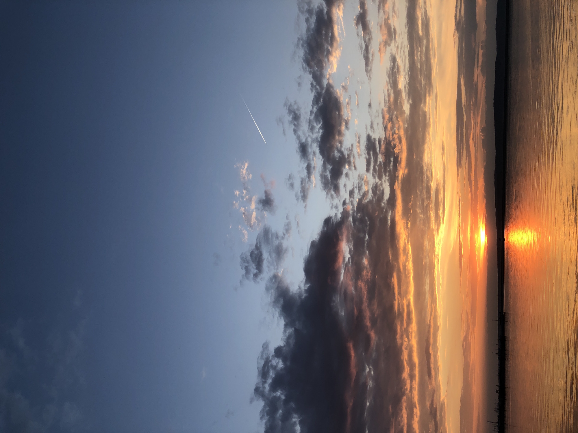 The image is of a sunset over water, potentially a large lake or river. The sky is divided into two main areas: a clear blue section on the left and a cloudy area on the right. The clouds are varying shades of gray and orange, illuminated by the setting sun. A contrail or shooting star is visible in the blue portion of the sky. The water reflects the colors of the sunset, with a bright orange glare where the sun meets the horizon. A dark, indeterminate structure or landmass is visible on the horizon, separating the water from the sky. The image appears to be rotated 90 degrees counterclockwise. Without additional information, identifying the location as Chaikovsky, Russia, is speculative.
