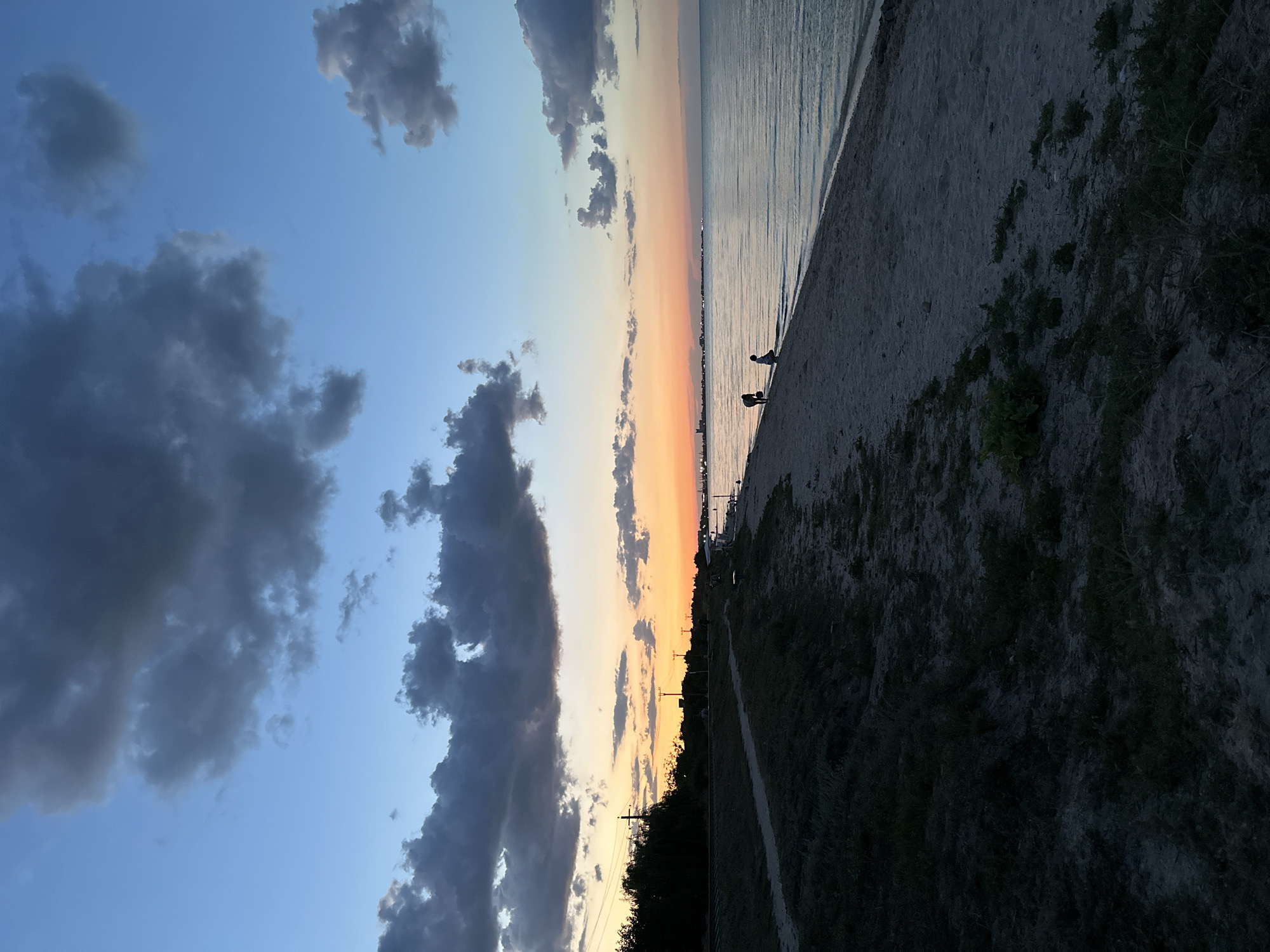 The image depicts a twilight scene on a beach, likely in Yeysk, Russia, given the context of its location. The sky is a gradient of blue and orange, transitioning from a deep blue at the top to a vibrant orange near the horizon, indicative of sunset. Several dark, fluffy clouds are scattered across the sky.
The foreground shows a sandy beach with sparse green vegetation growing along the edge of a grassy embankment. A dirt path runs parallel to the beach, separating the embankment from the sand. In the distance, along the shoreline, several people are visible. Two figures, appearing to be a child and an adult, are standing near the water's edge, possibly engaging in activities like playing or exploring. Further down the beach, other individuals can be seen, and a few benches are present.
Beyond the beach, the calm sea stretches towards a distant cityscape. The cityscape is silhouetted against the bright horizon, with several buildings and structures visible. Power lines and utility poles are also present along the edge of the land, contributing to the urban fringe feel of the location. The overall impression is of a serene, end-of-day scene at a coastal area.