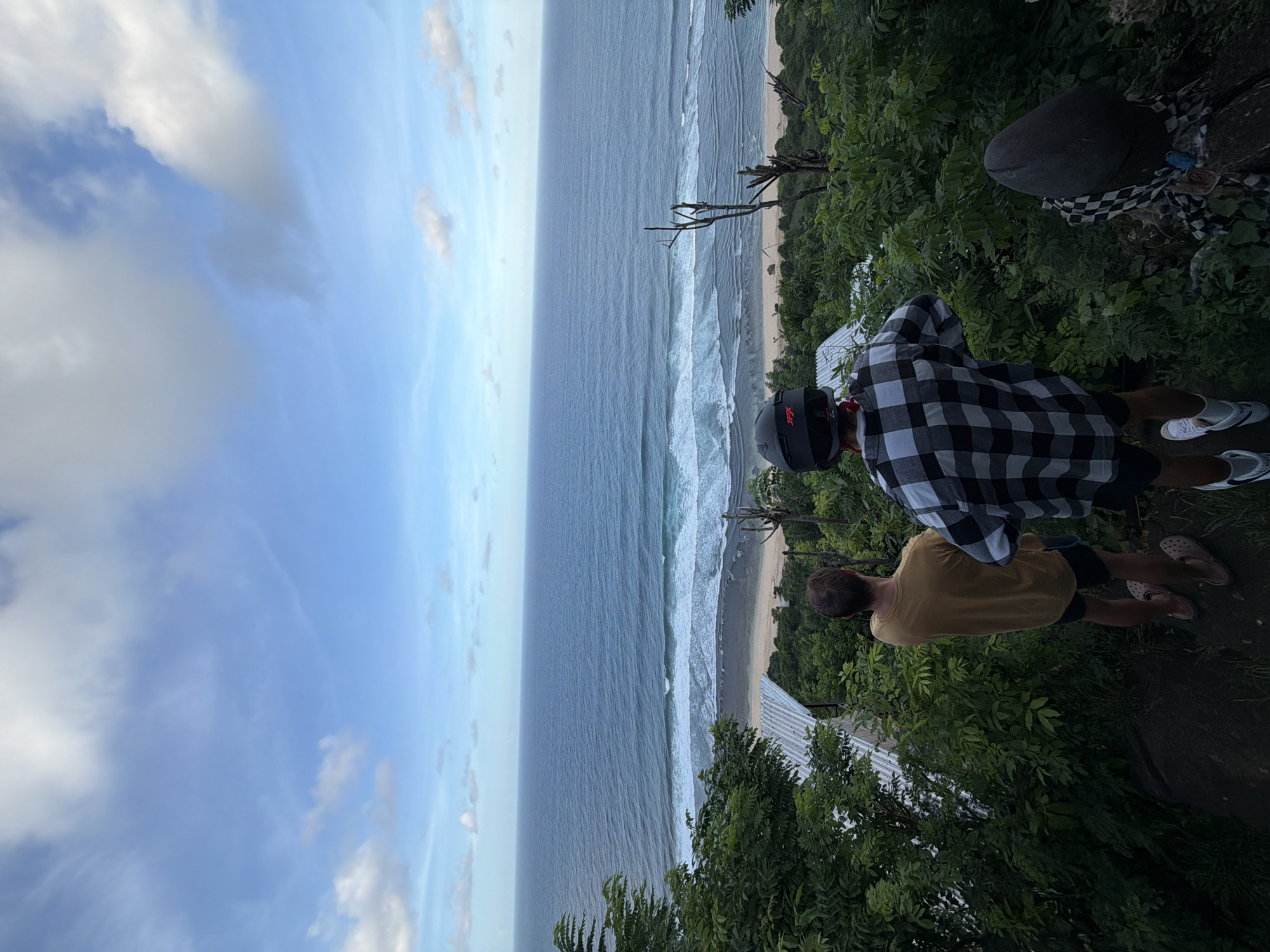 The image shows a scenic overlook in Kuta Selatan, Indonesia. The view encompasses a sandy beach, the ocean with visible waves, and a blue sky with clouds. Two people are present in the foreground, standing near the edge of the overlook. One is wearing a plaid shirt and a helmet, and the other is wearing a yellow shirt. Both appear to be looking out at the view. Green vegetation surrounds the overlook.