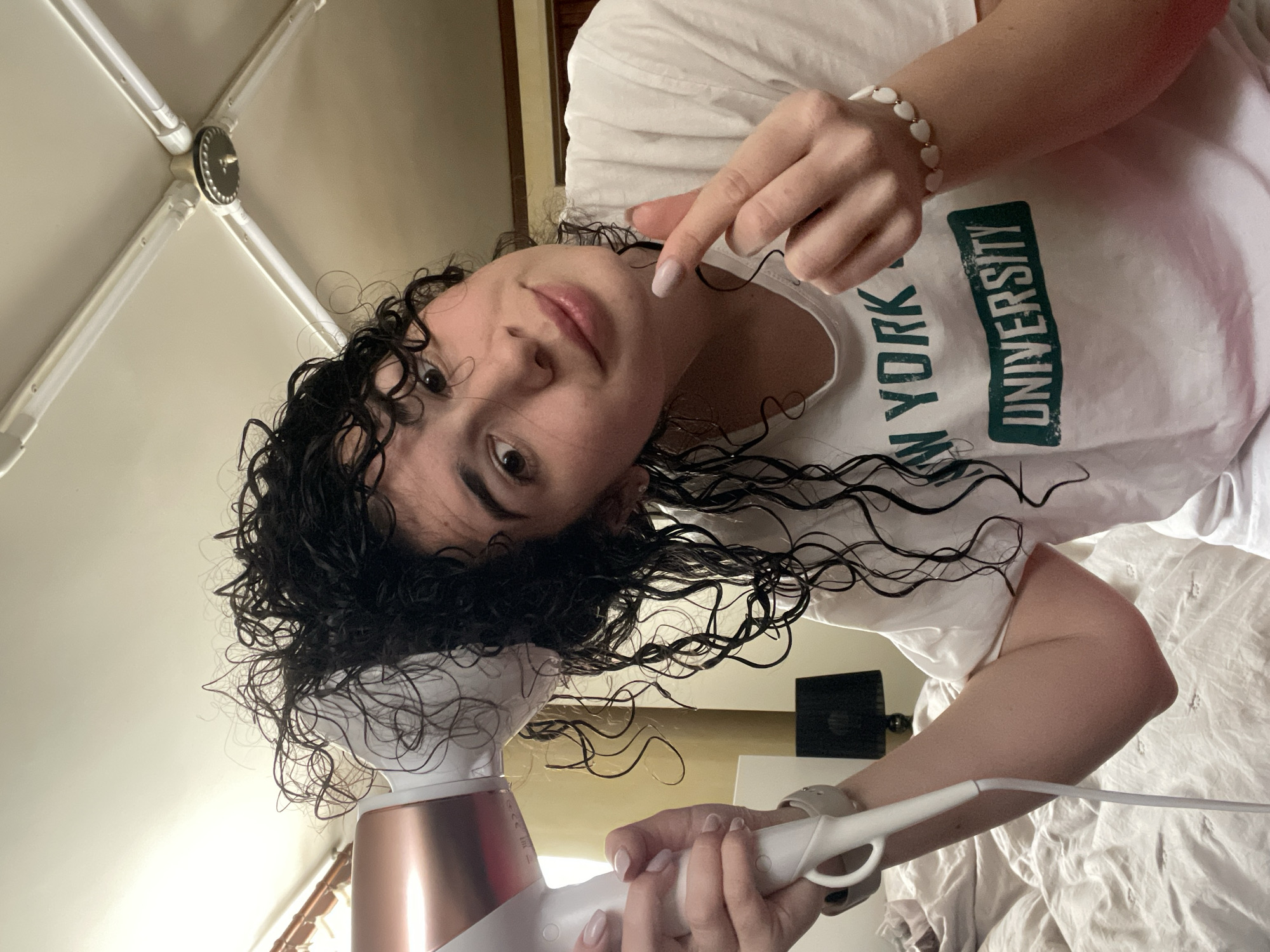 This media file captures a low-angle, rotated view of a young person in an indoor setting, actively drying their wet, curly hair with a hairdryer.
The central subject is a young person with light skin, dark eyes, and abundant, dark, wet, curly hair that forms distinct ringlets. They are looking towards the camera with a soft, slightly smiling expression. They wear a simple white t-shirt featuring the text "NEW YORK UNIVERSITY" printed vertically in bold green letters. On their right wrist, they wear a white beaded bracelet with small golden heart accents. Their fingernails are painted with a light, possibly pale pink or nude, polish.
The person is holding a hairdryer, predominantly white with rose gold or copper-toned accents, in their left hand, directing it towards their wet hair. Their right hand is raised, with the index finger extended, pointing upwards.
The setting appears to be a personal space, likely a bedroom. White, somewhat rumpled bedding is visible in the lower right portion of the image. Above the person's head, a white ceiling structure with a central dark, circular fixture is discernible, possibly a light fixture or part of an architectural element. No specific visual cues confirm the given location of Sofia, Bulgaria. The time of day is not explicitly indicated, but the lighting suggests an indoor, well-lit environment. The overall mood is casual and candid.